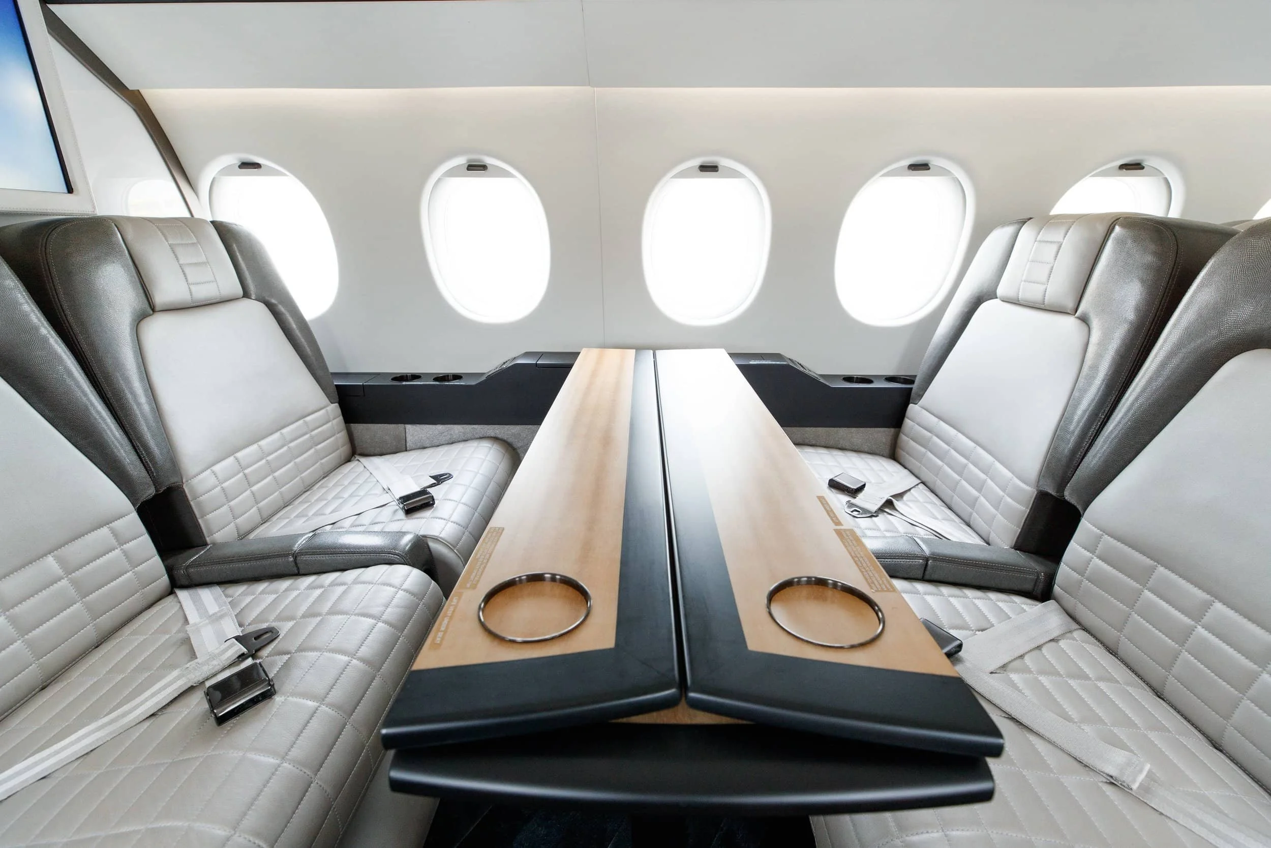 Empty airplane business class cabin with two rows of cream and gray leather seats, sharing a central table, with six windows visible in the background.