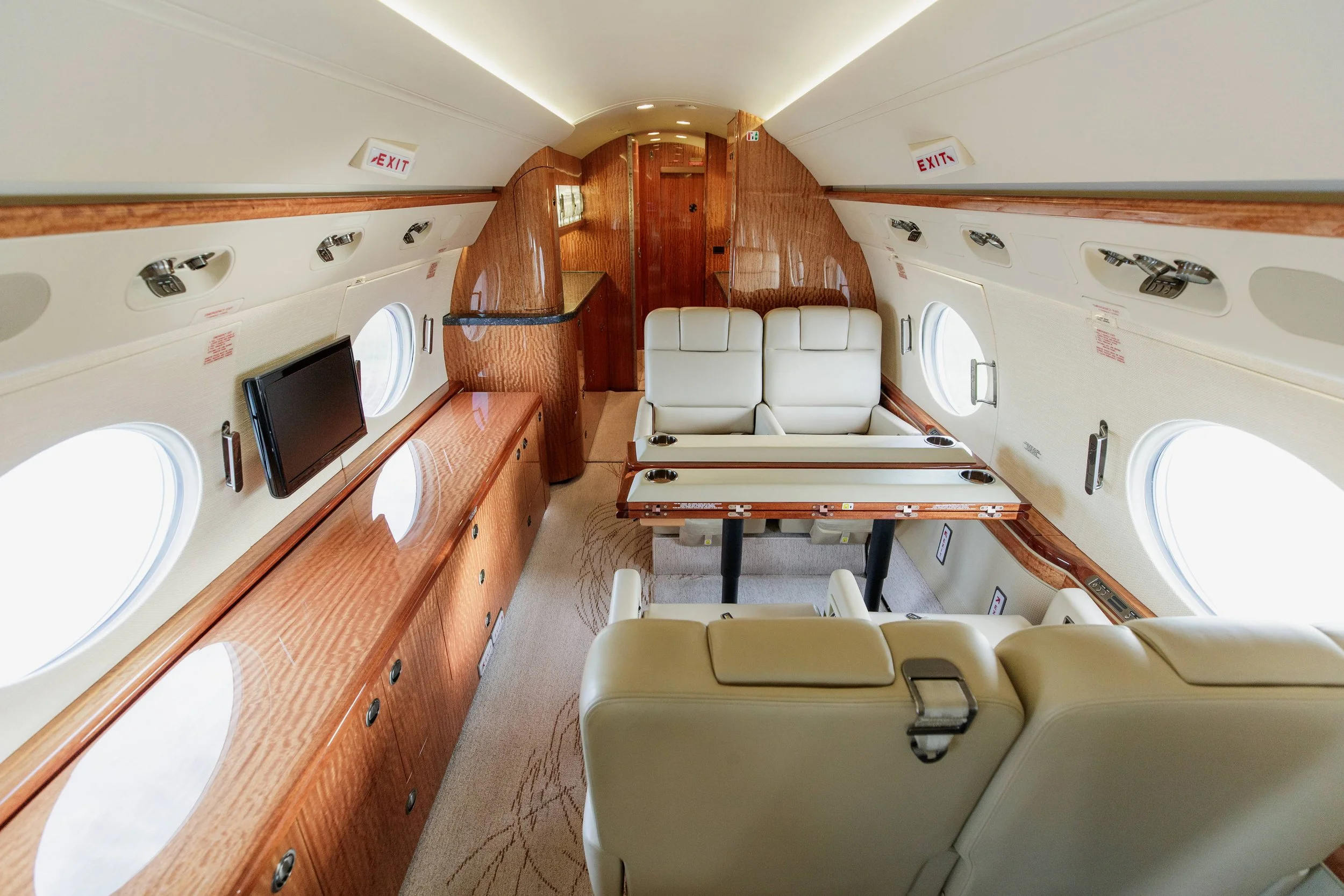 Interior of a private jet with cream leather seats, wood paneling, television, and multiple windows.