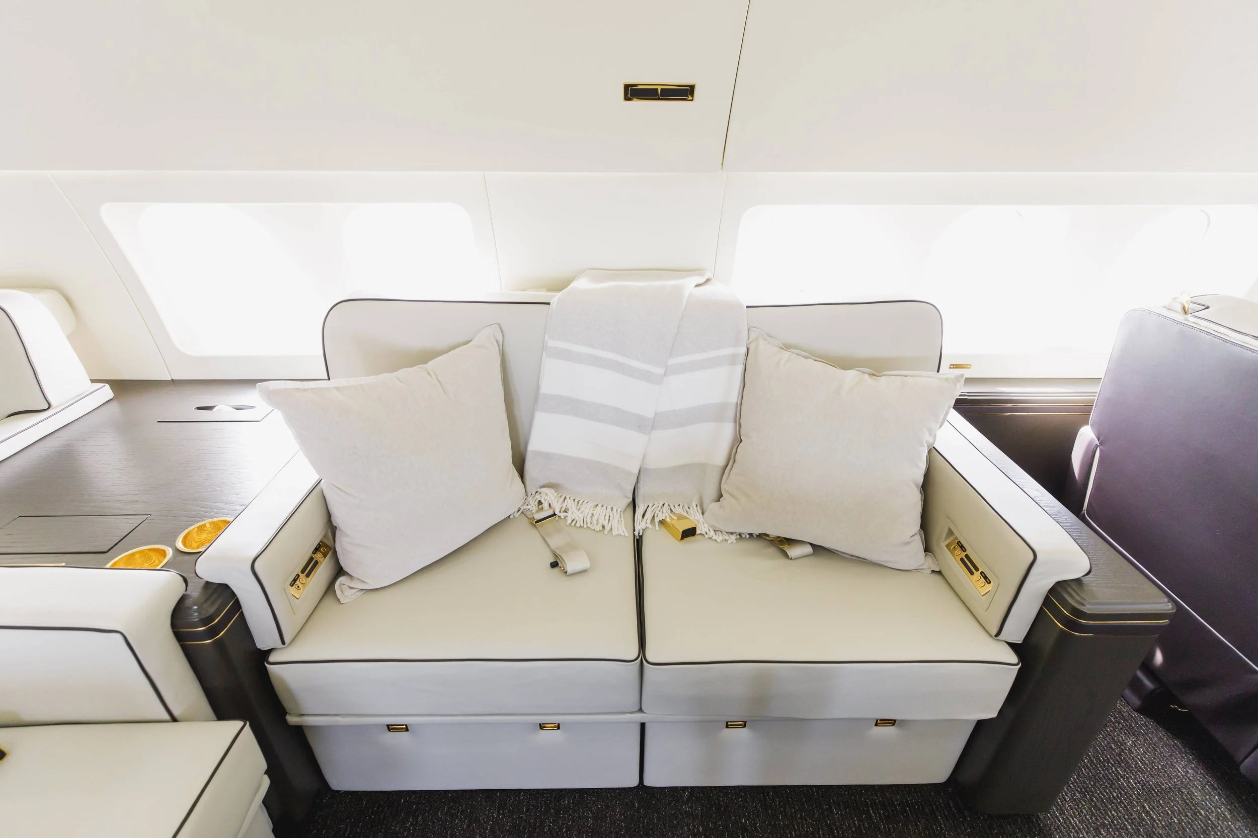 Luxury airplane seat with beige cushions and a striped towel draped over the headrest, next to a side table with gold accents.