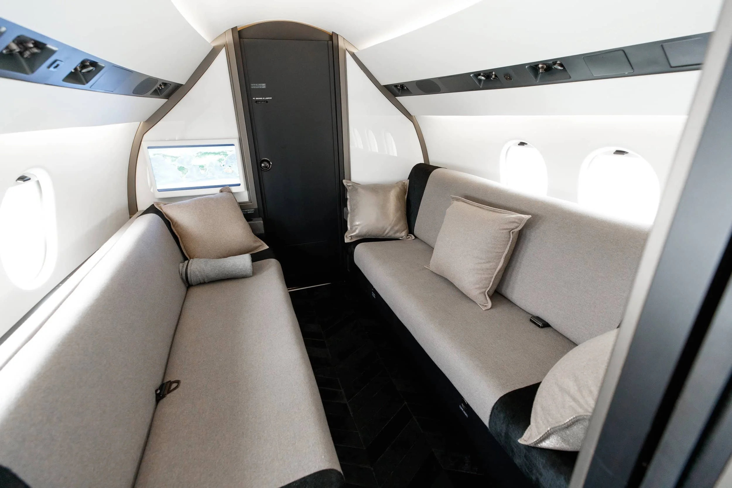 Inside the cabin of an airplane, showing two beige upholstered seats with pillows and a window on the left side.