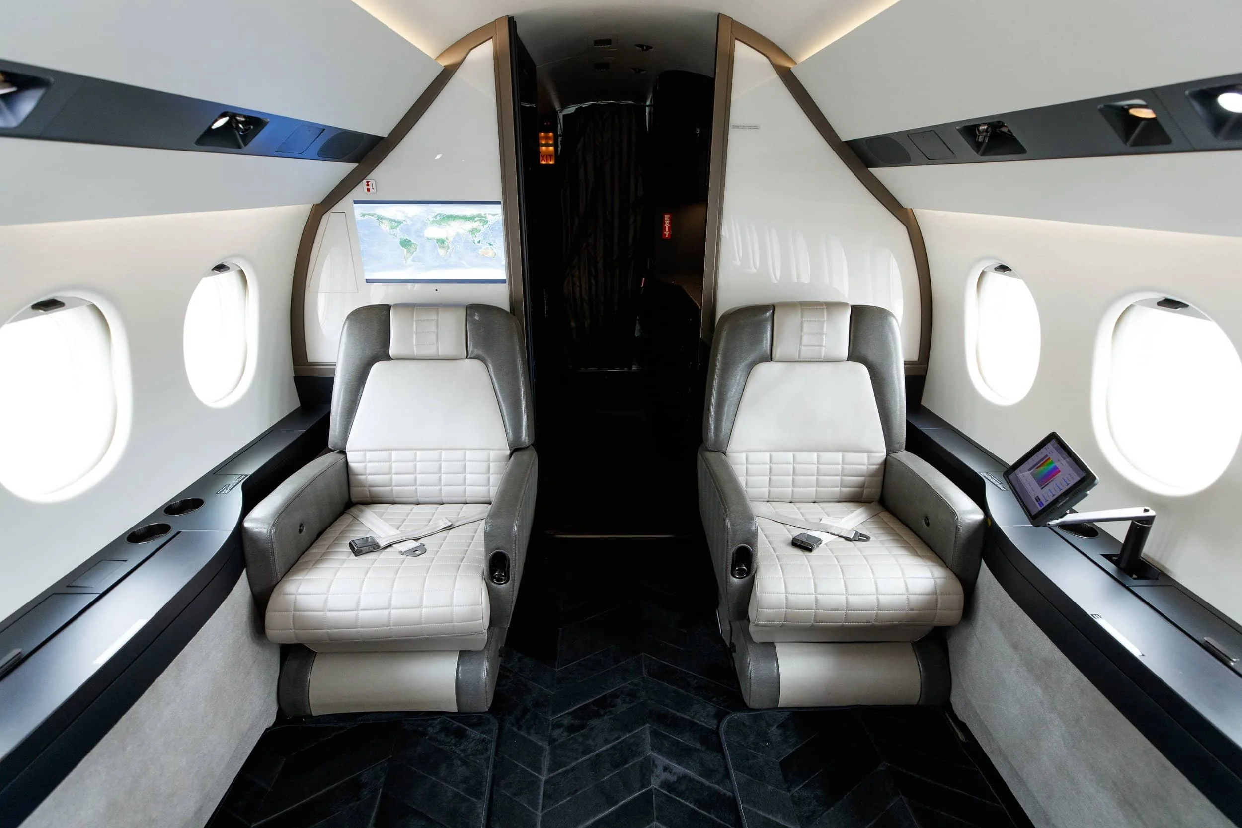 Interior of a private jet with two upholstered seats facing forward, each with seat belts, multiple windows along the walls, a small screen on the right side, and a window showing a world map.