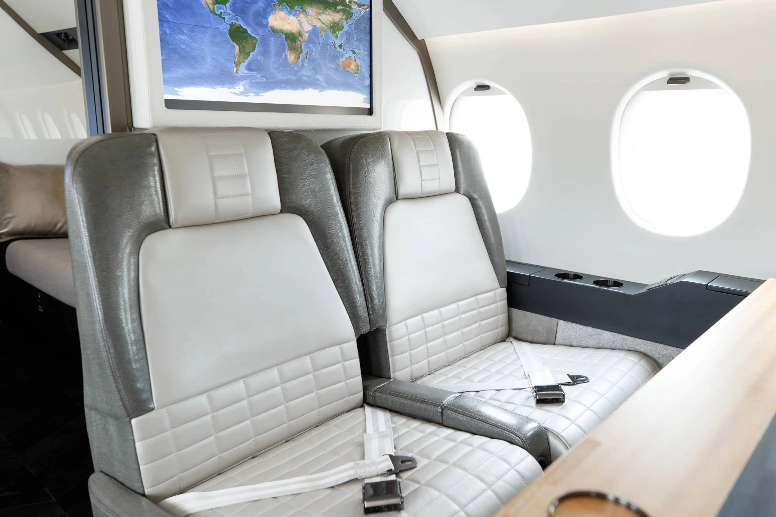 Inside an airplane cabin showing two beige and gray leather seats with seat belts, a window, and a mounted screen showing a world map.