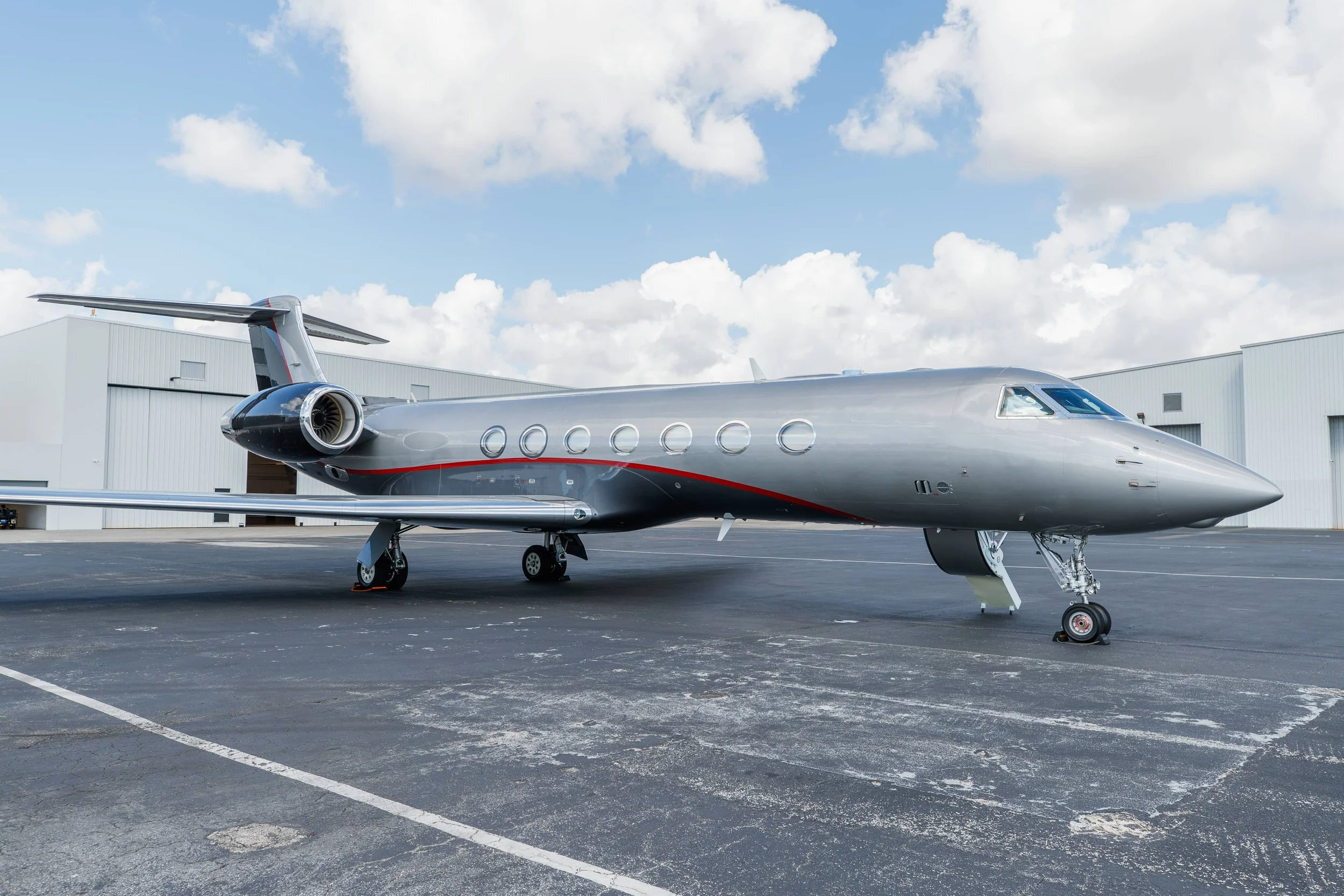 A sleek, silver private jet parked on an airport tarmac with hangars in the background and partly cloudy skies above.