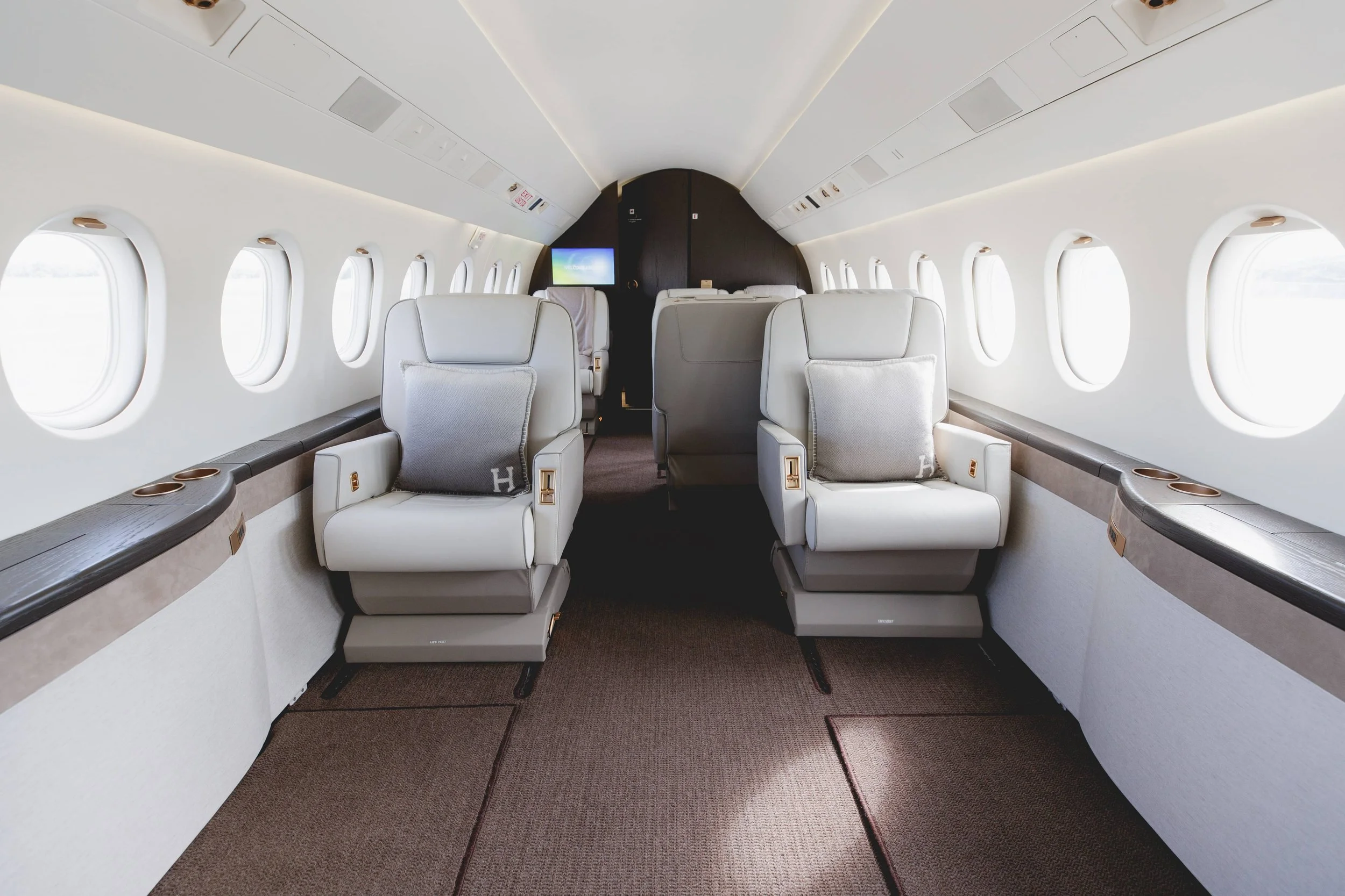 Interior of a private jet with two white leather seats facing forward and large oval windows on both sides, some small cushions on the seats, and a small screen at the front.
