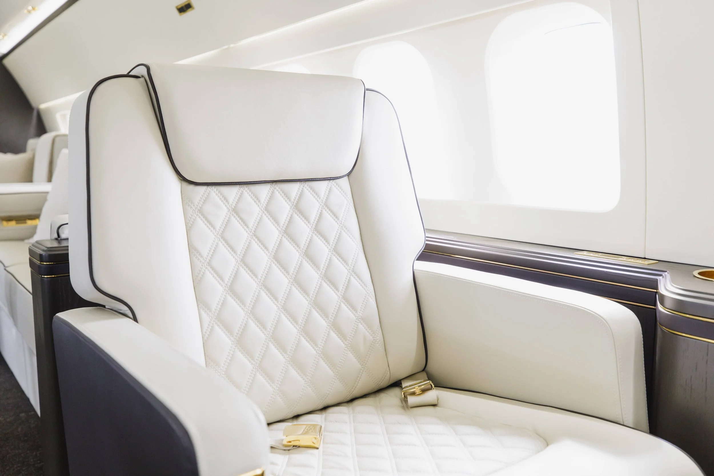 Interior of a luxury airplane with a white leather seat featuring quilted detailing, a black piping trim, and large windows.