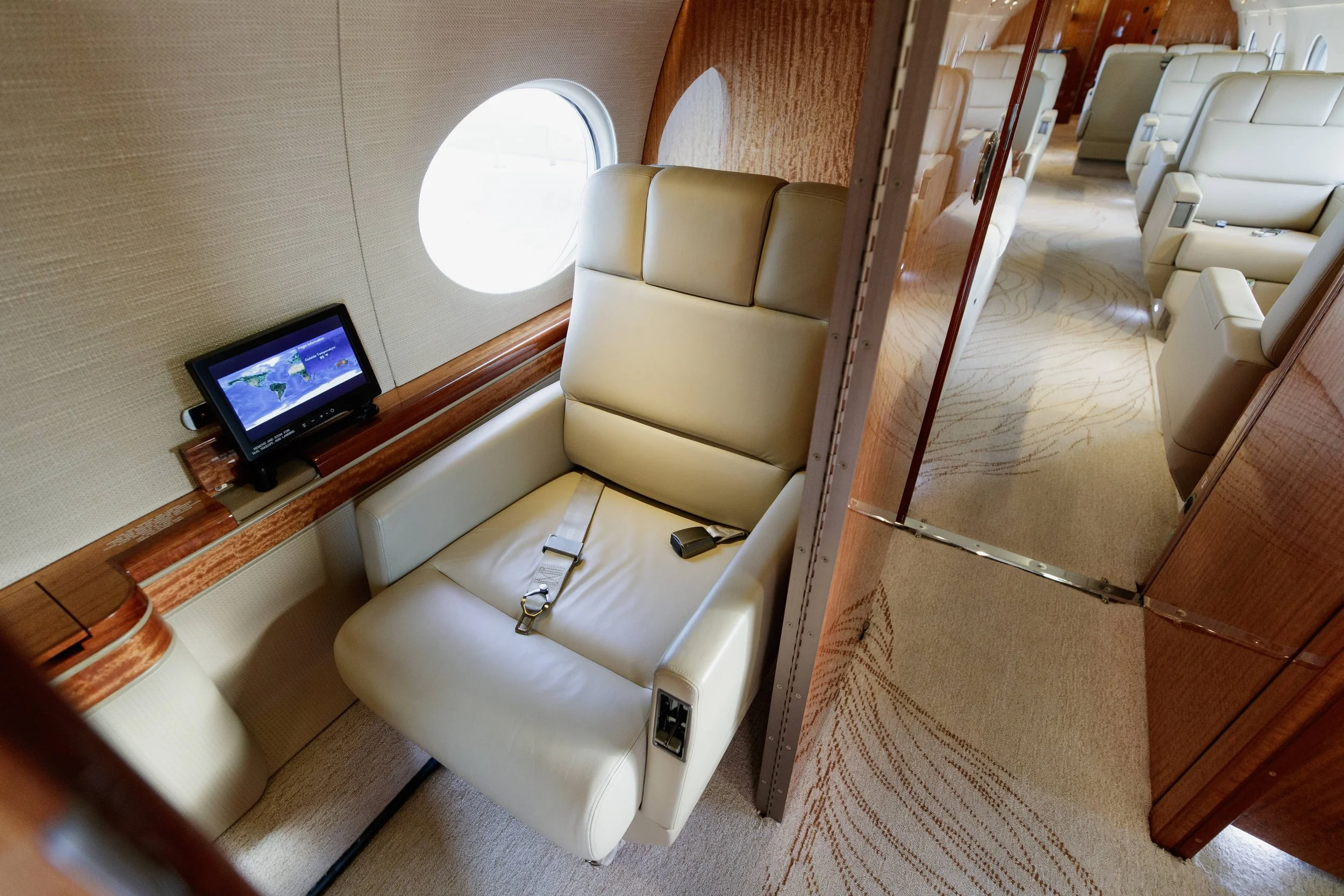 Interior of a private jet with beige leather seats, a window, and a small screen displaying a world map.
