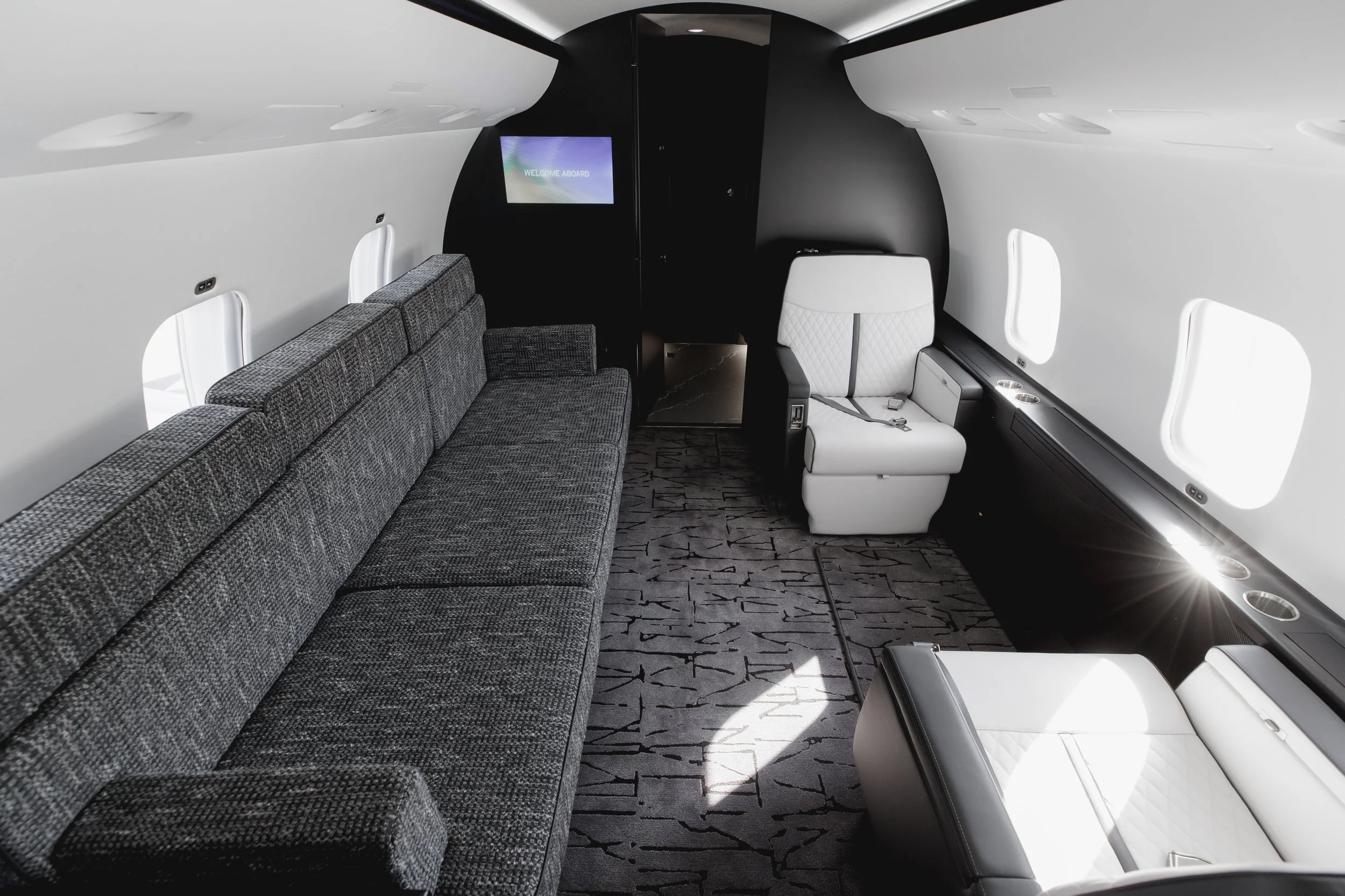 Interior of an airplane cabin with seating area, including a long gray upholstered sofa on the left and white leather seats on the right. There are windows along the side walls, and a screen on the back wall displaying "WELCOME ABOARD."