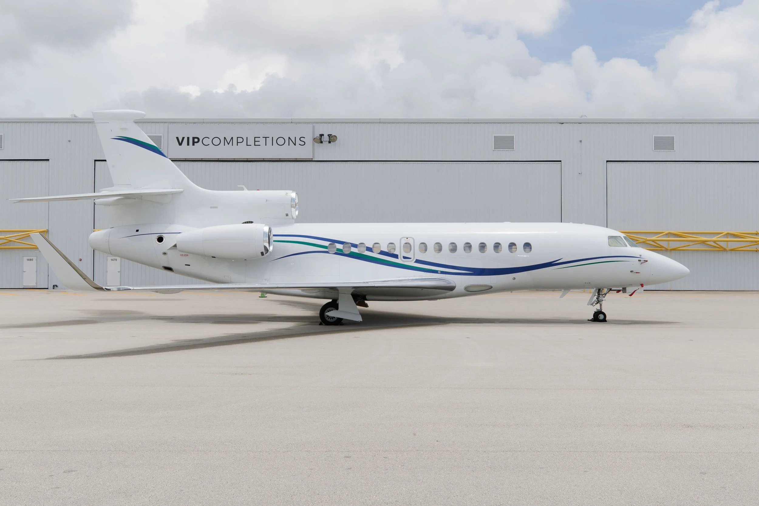 White private jet aircraft parked on tarmac in front of a building labeled VIP COMpletions, with cloudy sky overhead.