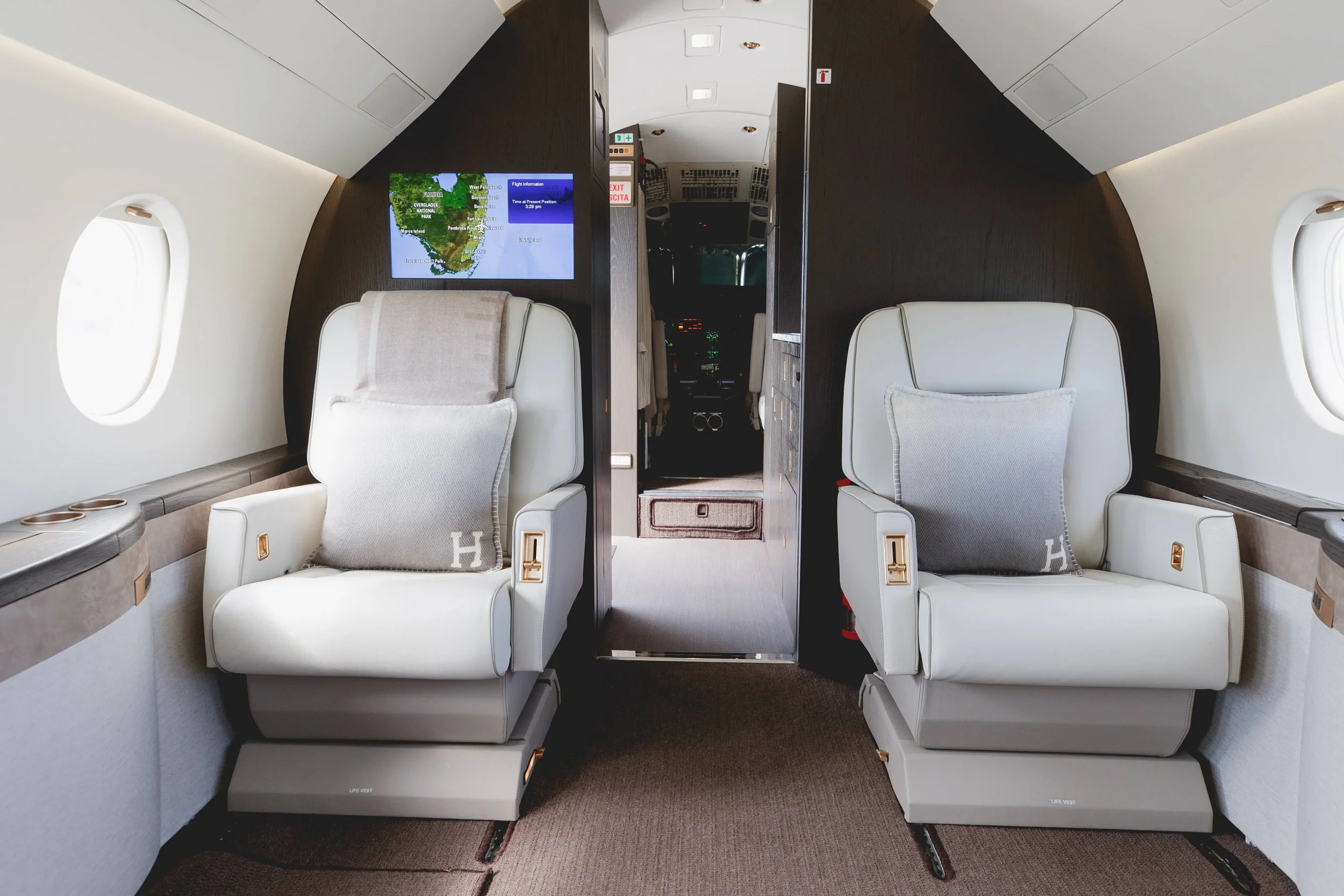 Two plush white airplane seats with gray pillows inside an aircraft's cabin, positioned next to oval windows, with a small TV screen displaying a map and flight information on the wall between the seats and the cockpit in the background.