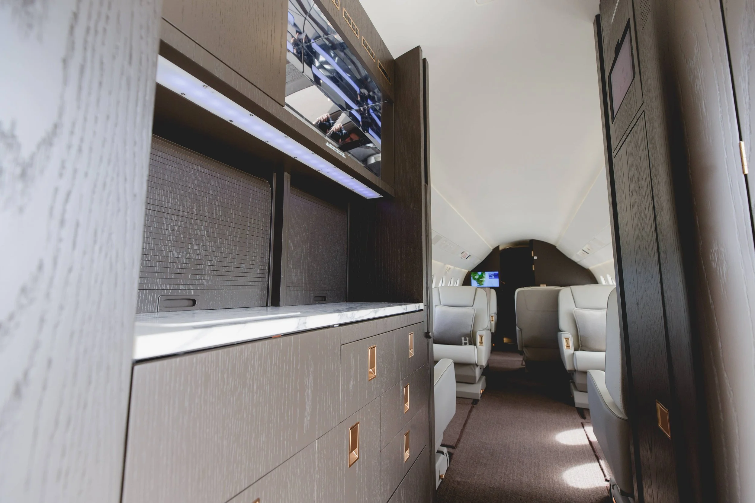 Interior of a modern private jet with beige leather seats, dark wood paneling, and a television screen on the wall.