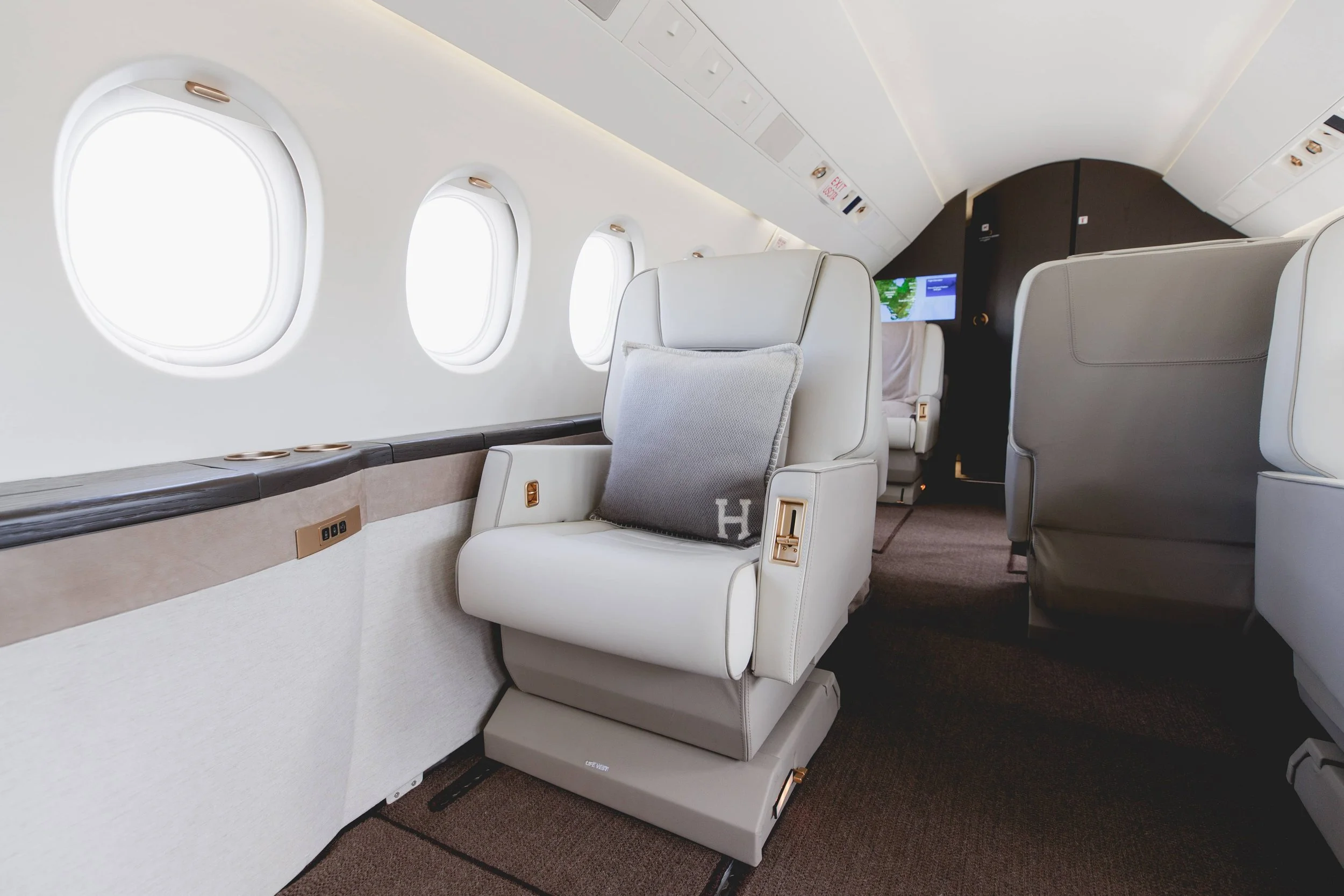 Luxury airplane cabin interior with plush beige seats, one with a gray pillow, and three view windows on the left side. The cabin has a modern design with dark and light accents.