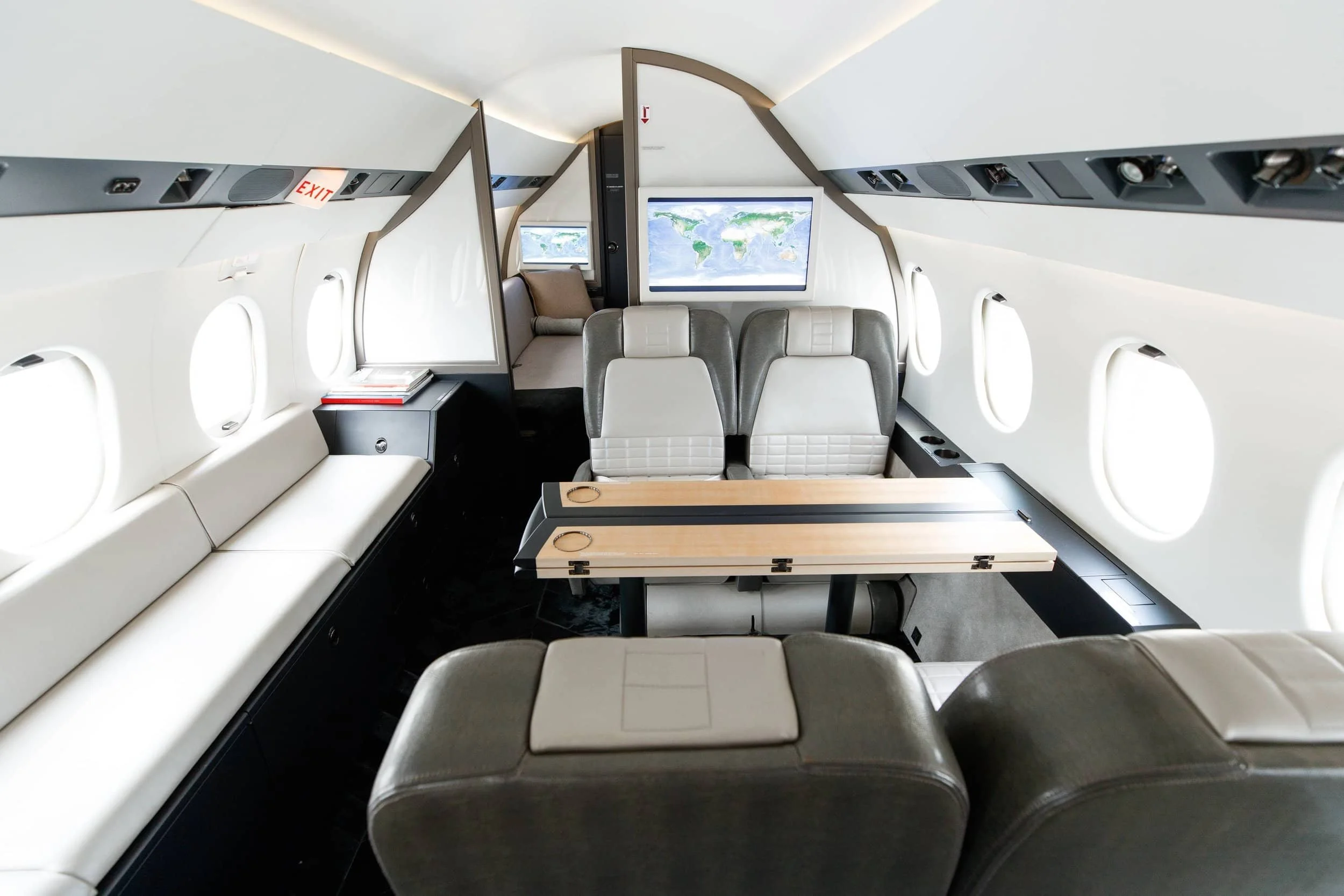 Interior of a private jet with beige and gray seats, a folding table, and a large screen displaying a world map in front of the seating area.