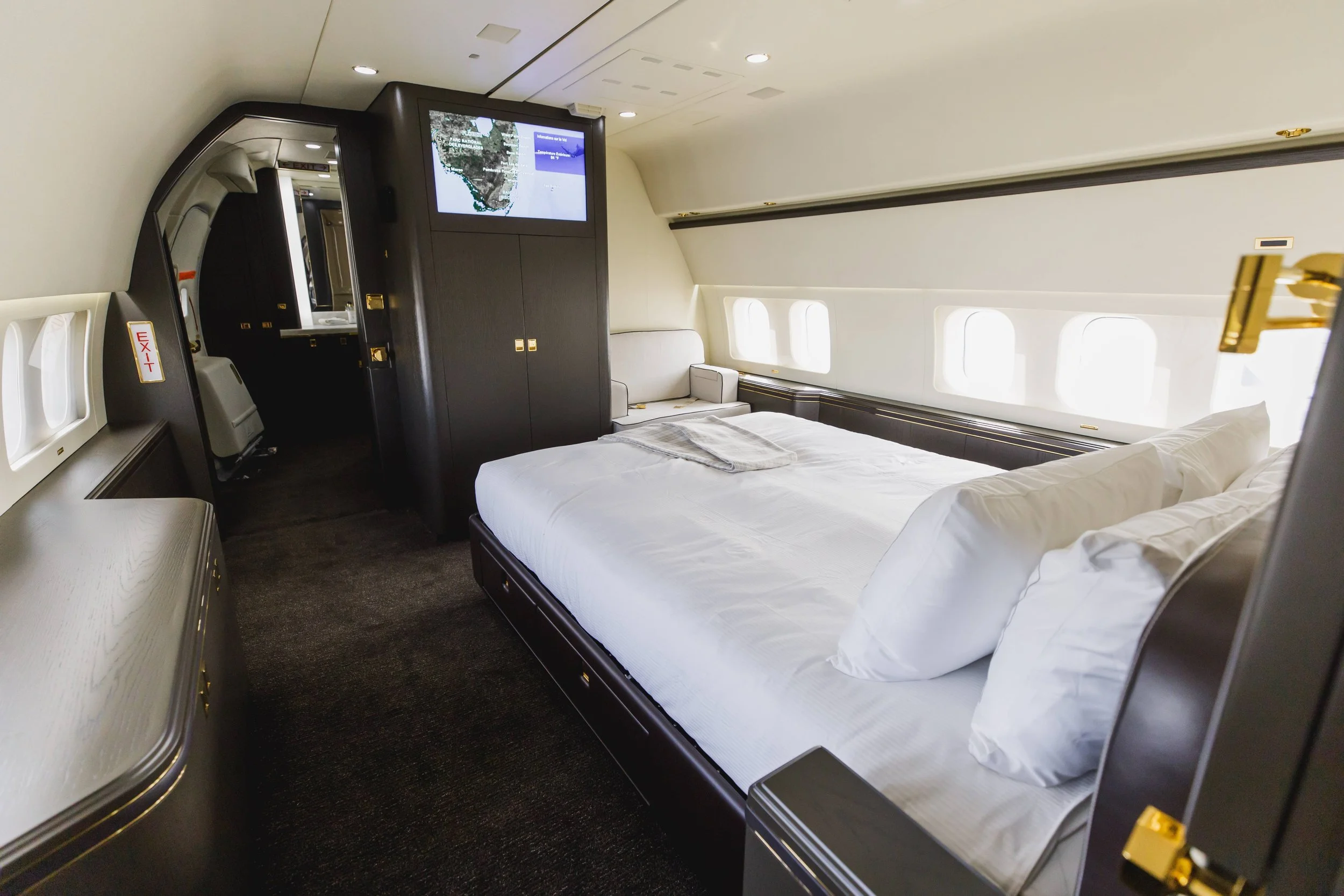 Luxury airplane bedroom with a bed, pillows, and a window-lined wall, featuring dark wood and white bedding.