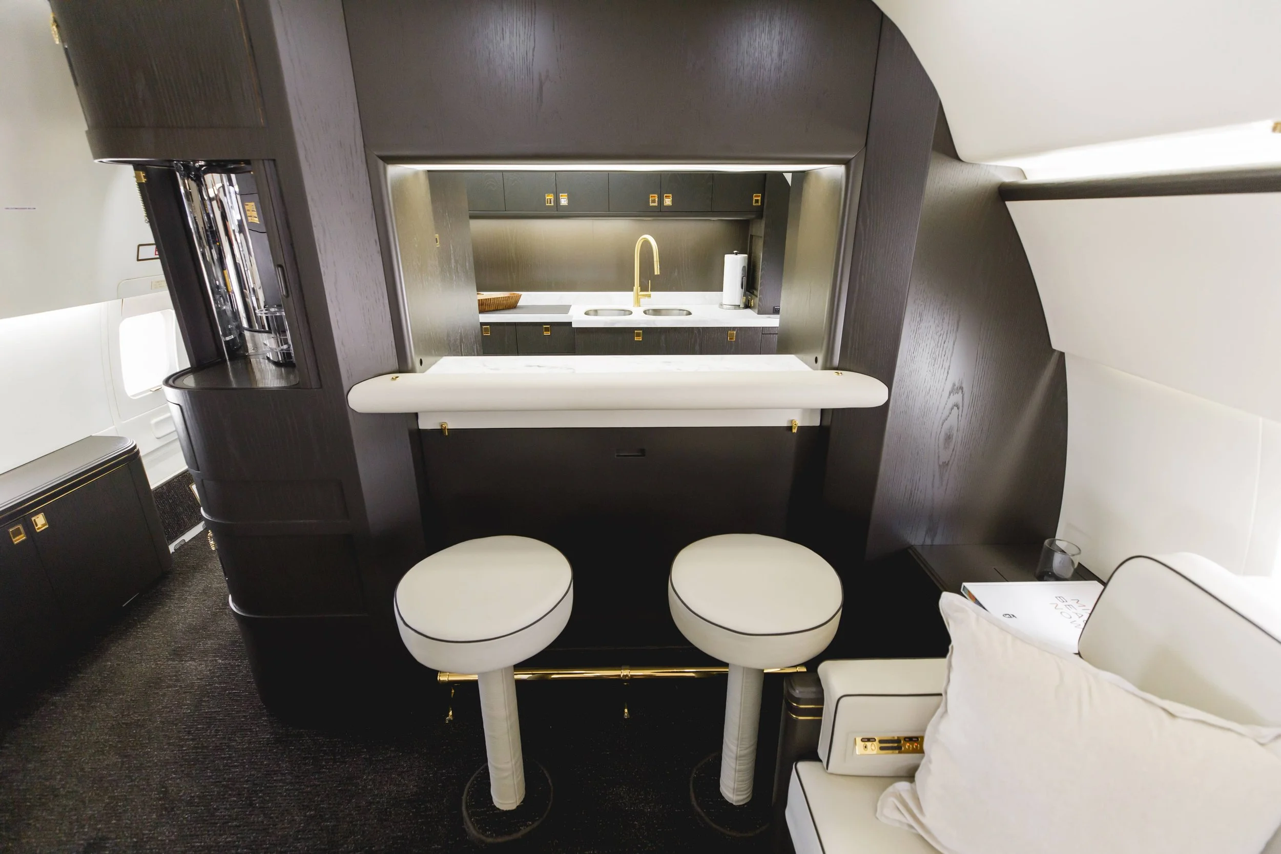 Interior of a luxury airplane cabin featuring a small kitchenette with a sink, gold faucet, dark cabinets, and a sitting area with two white stools and a white pillow on a seat.