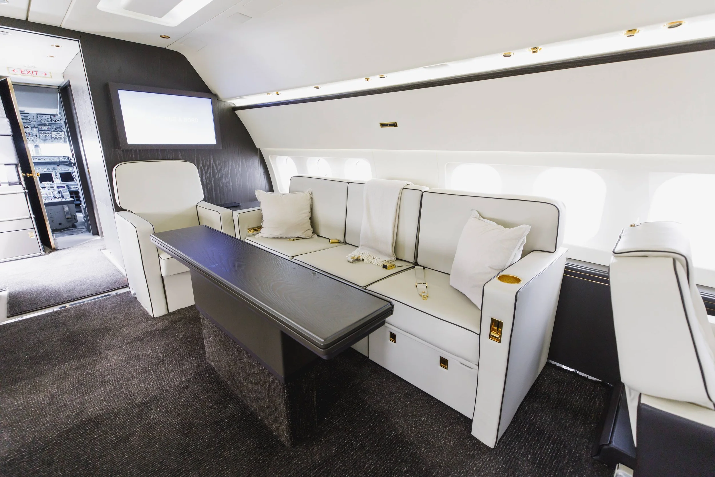 Interior of a private jet with white leather seating, a black wooden table, and a dark wood panel background. There are pillows on the seats, windows along the side, and an open door revealing the cockpit.