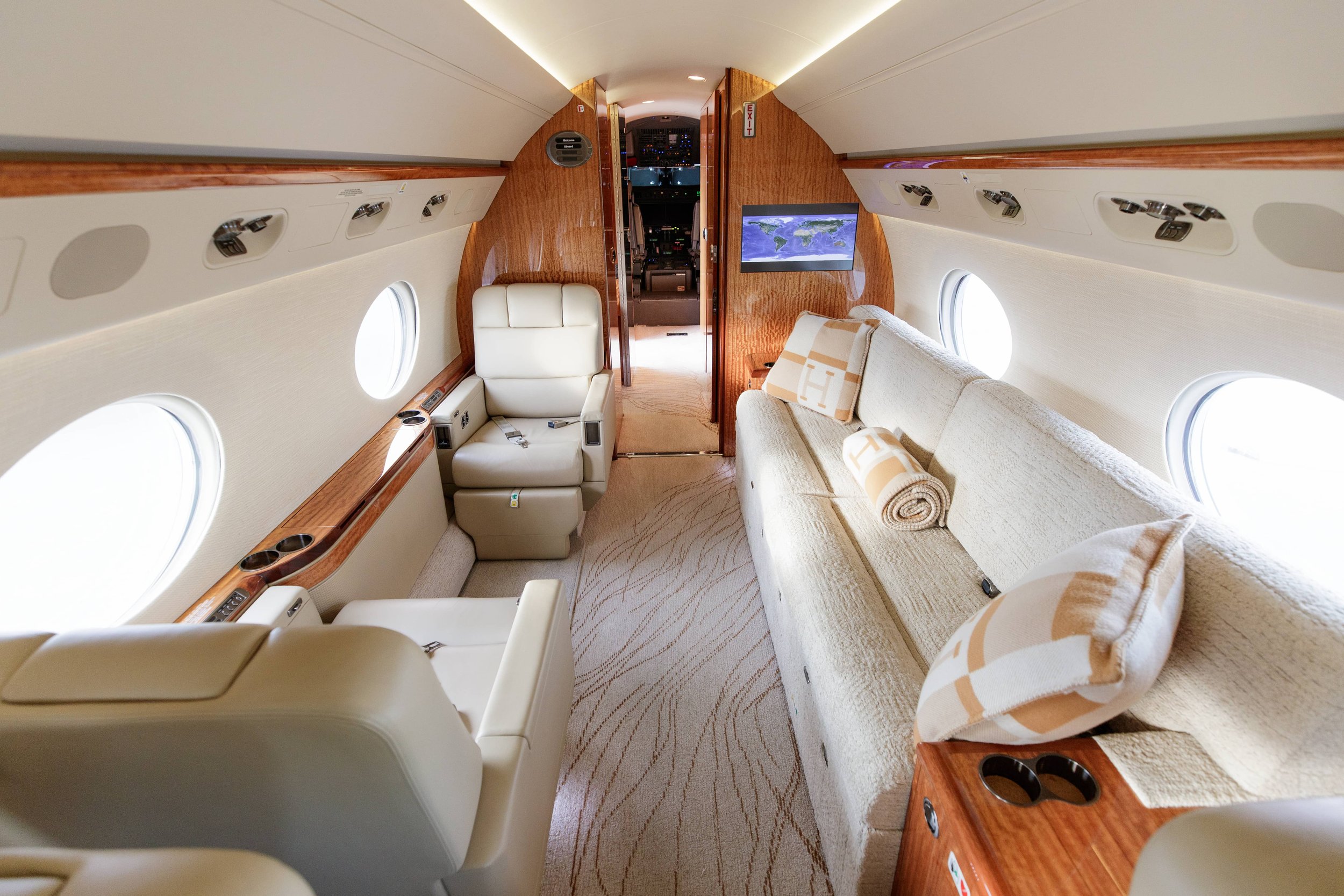 Interior of a luxury private jet with cream-colored seats and a couch, oval windows, wooden accents, and a TV screen.