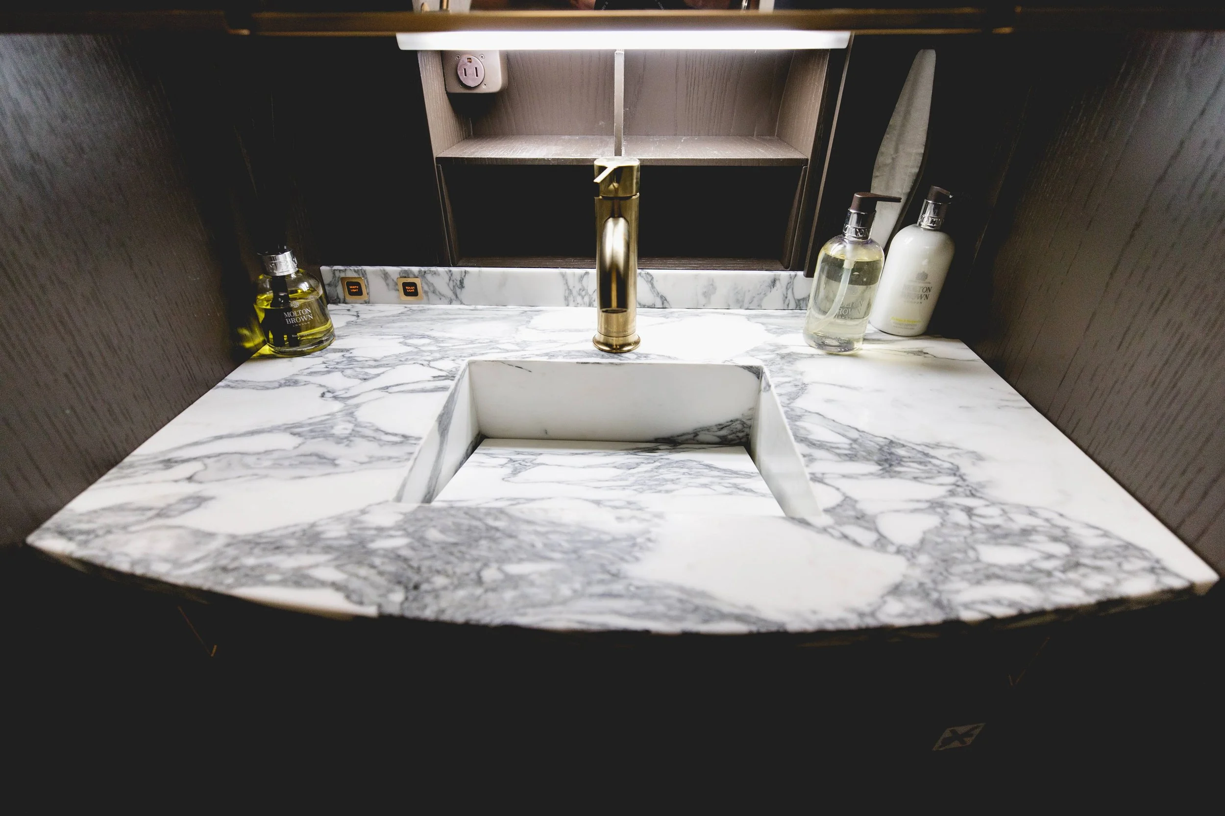 Bathroom sink with white marble countertop, gold faucet, and three bottles of soap or lotion on the right side. A mirror is mounted above the sink with a shelf above it, and a wall socket is visible on the back wall.