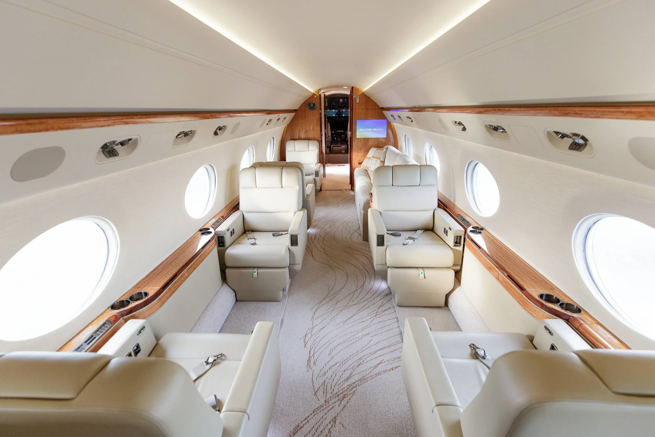 Interior of a private jet with cream leather seats, small round windows, and wood accents, facing forward with a view of the cockpit door at the front.