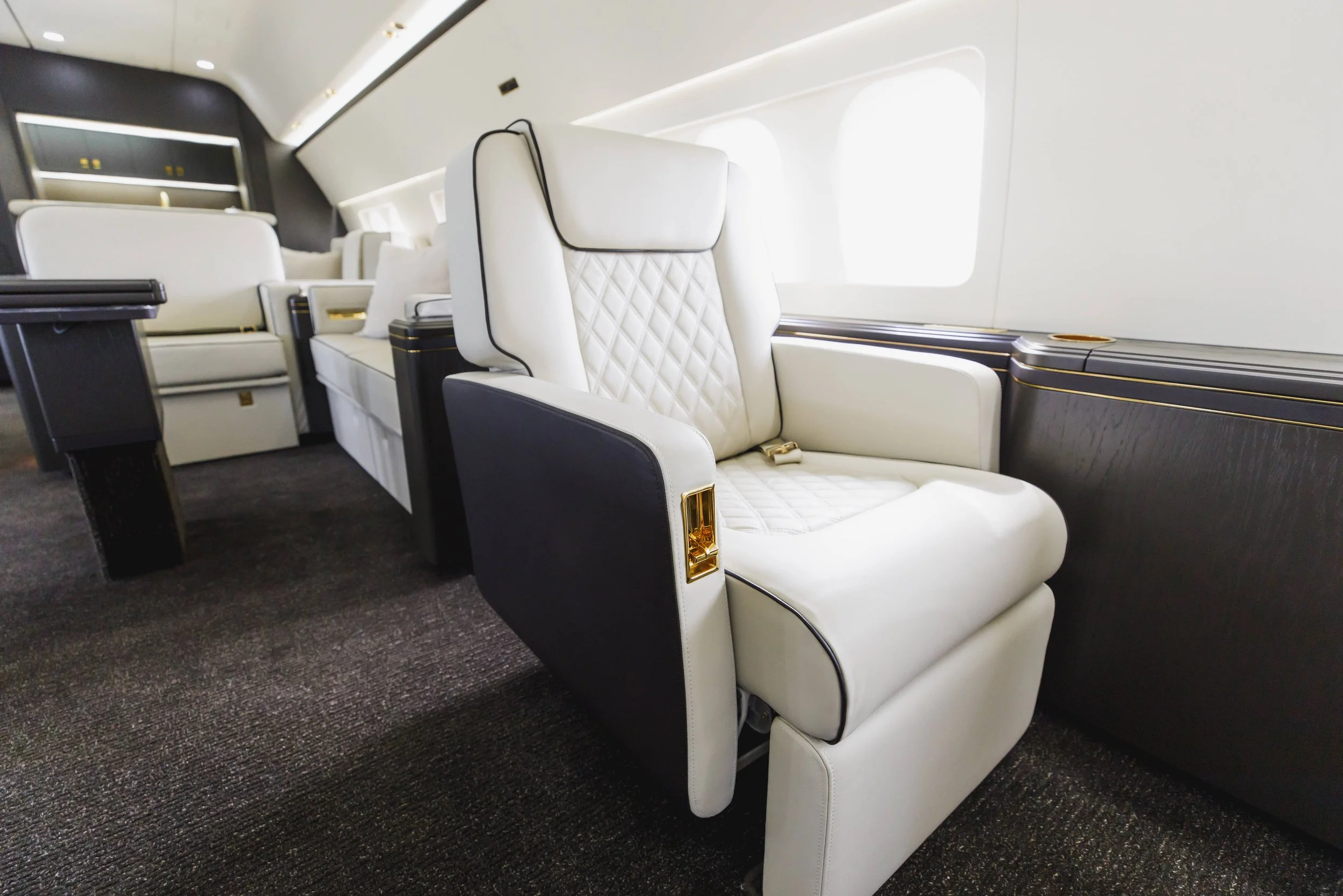 Luxury airplane interior with cream-colored leather seats and gold accents, showing a single seat near the window.