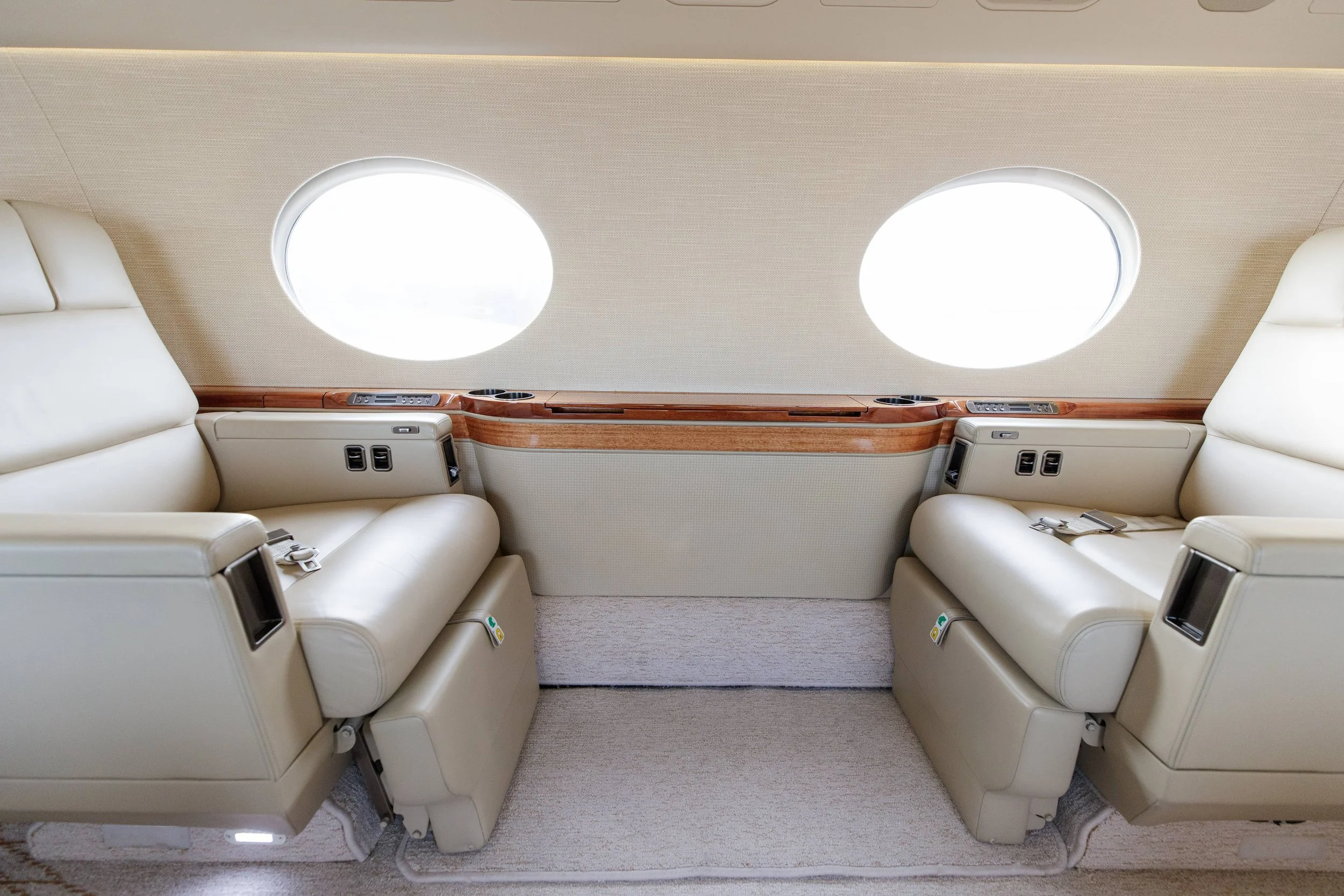 Interior of a private jet with two cream-colored leather seats facing each other, wooden side panels, and two circular airplane windows.