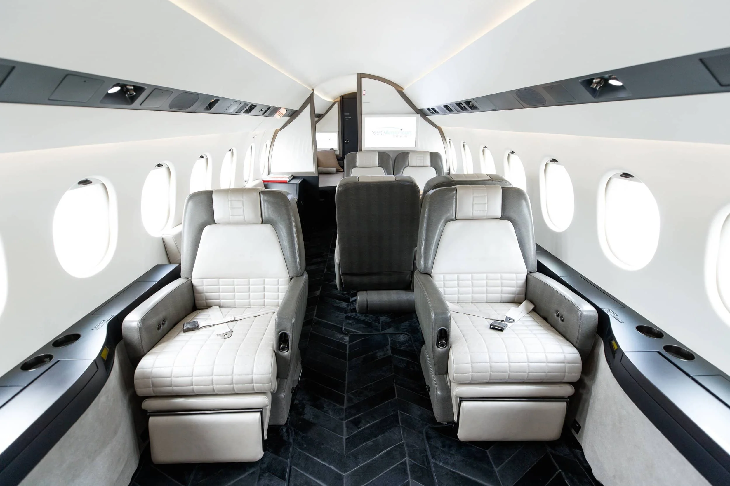 Inside a private jet, showing leather seats, windows along the side, and a screen at the front.