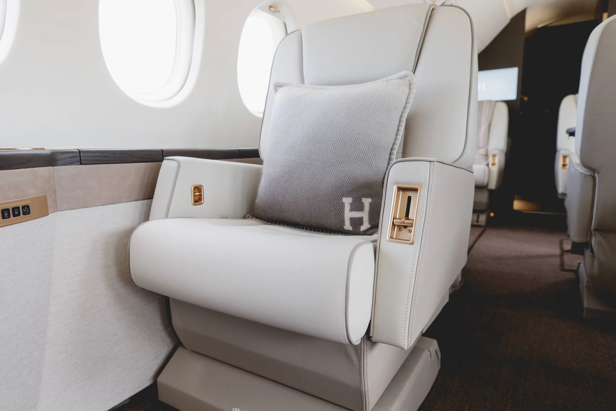 First class airplane seat with a grey cushion and a beige leather armrest, surrounded by other seats and windows.