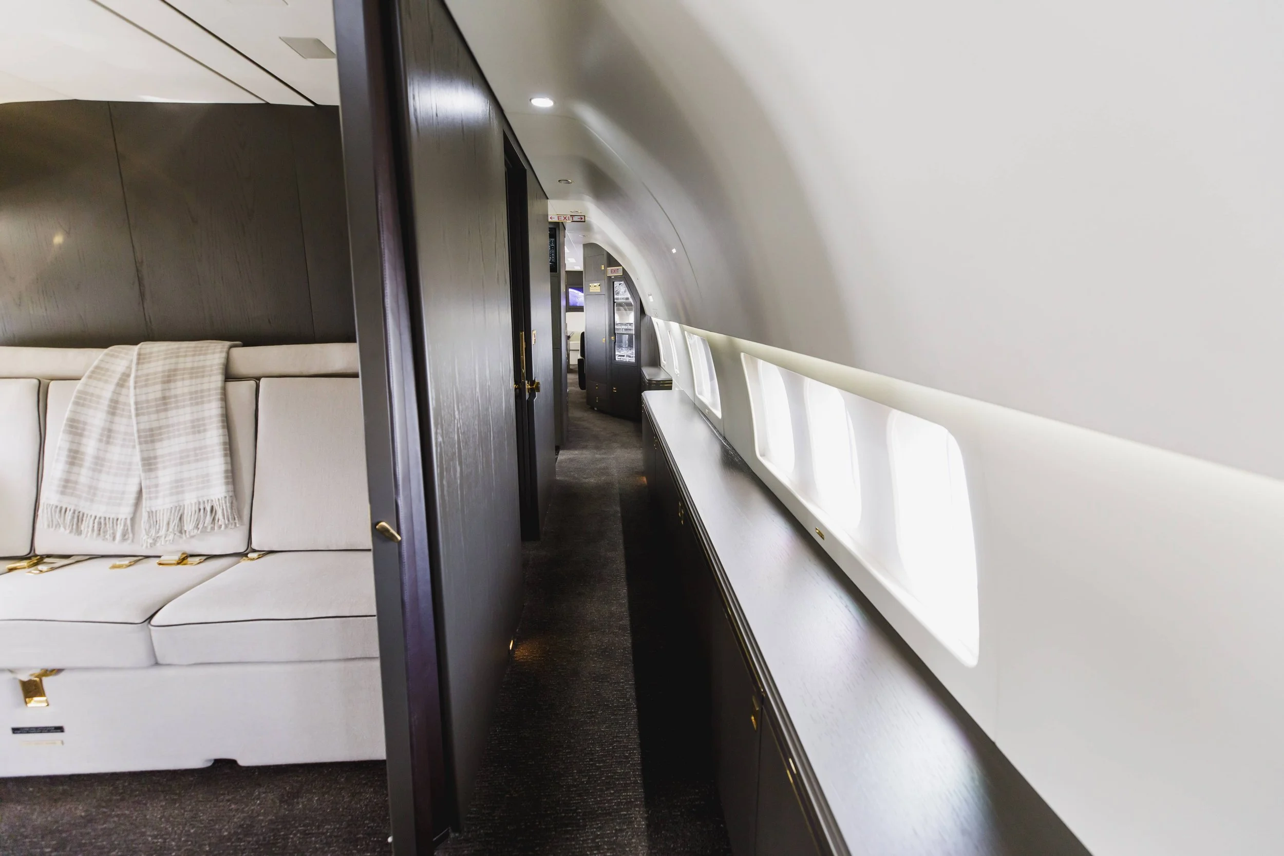 Interior of a private jet with a cream-colored sofa and a blanket, dark wood paneling, a narrow aisle, and windows along the side of the aircraft.
