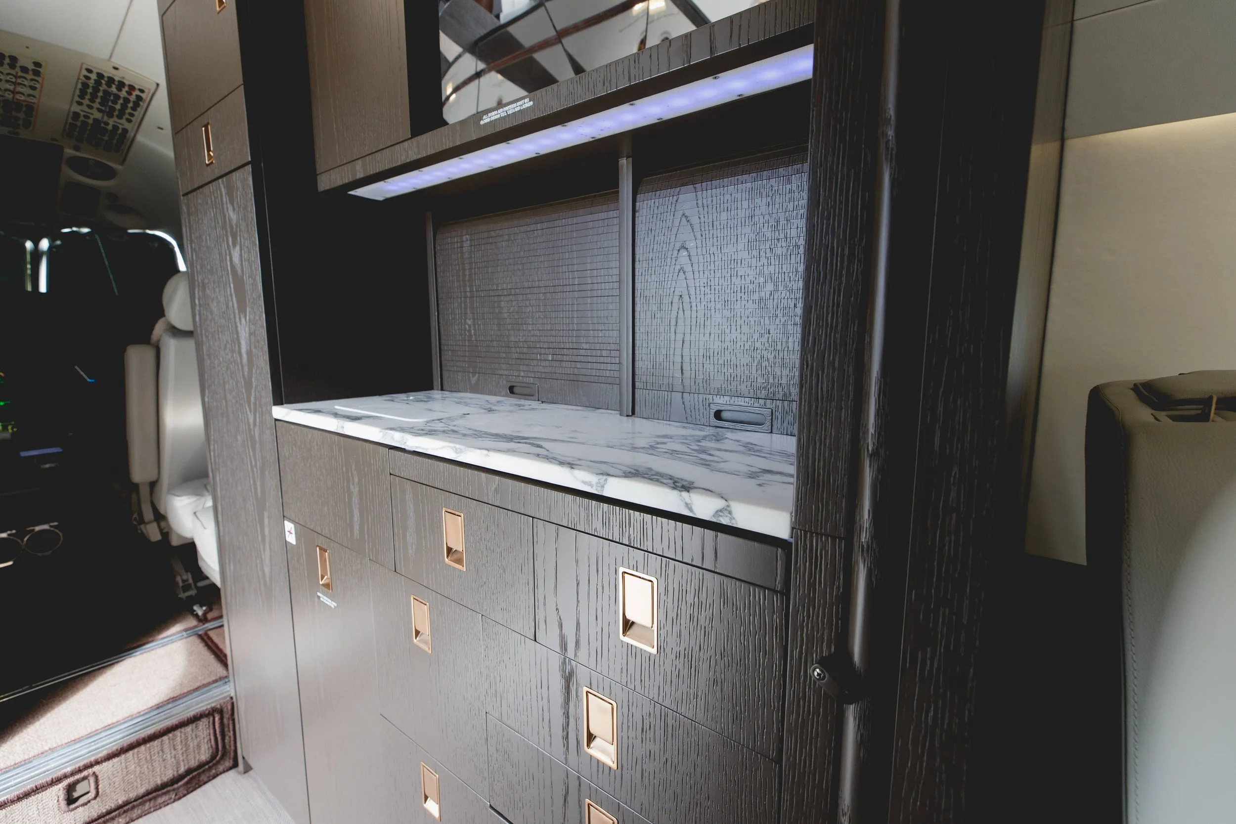 Inside a luxury jet, showing a dark wooden cabinet with gold handle accents and a marble countertop, with beige leather seats nearby.