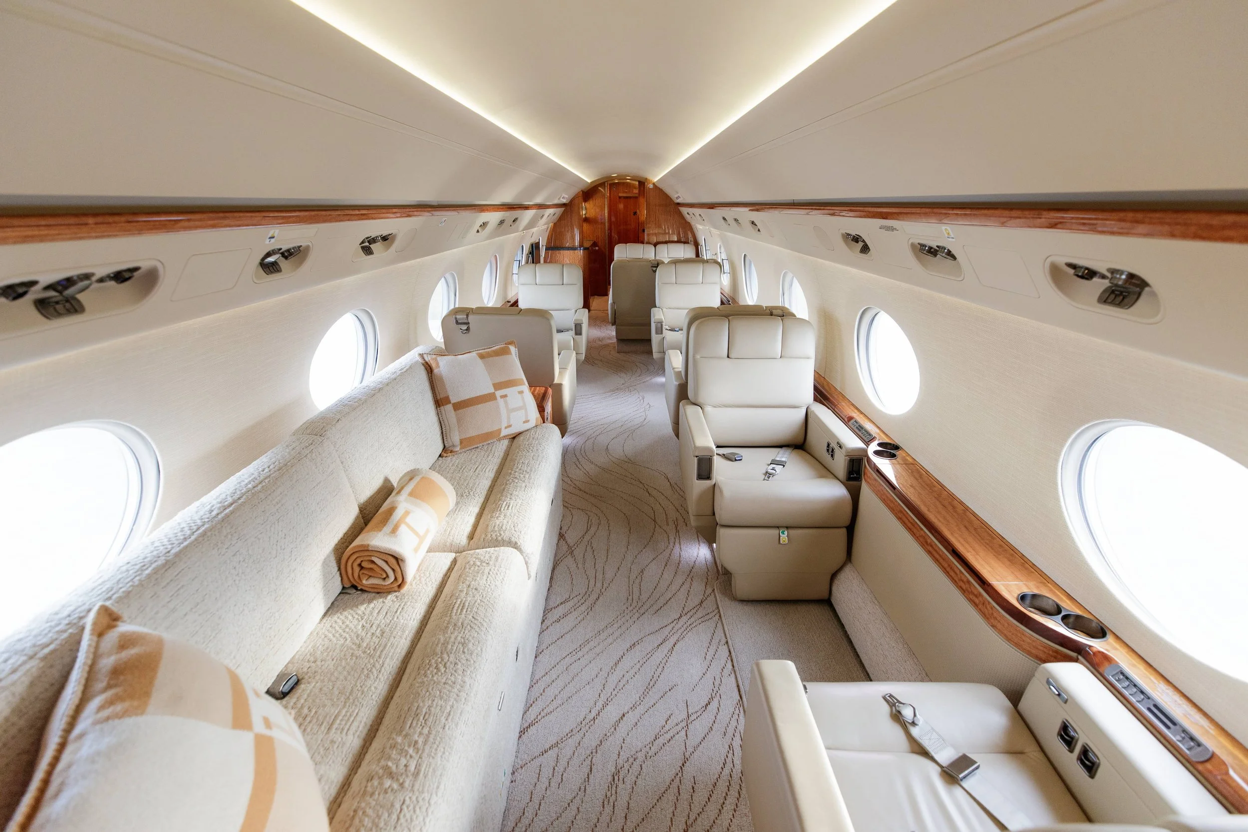 Interior of an airplane with white leather seats, beige carpet, round windows, and wooden paneling on the walls.