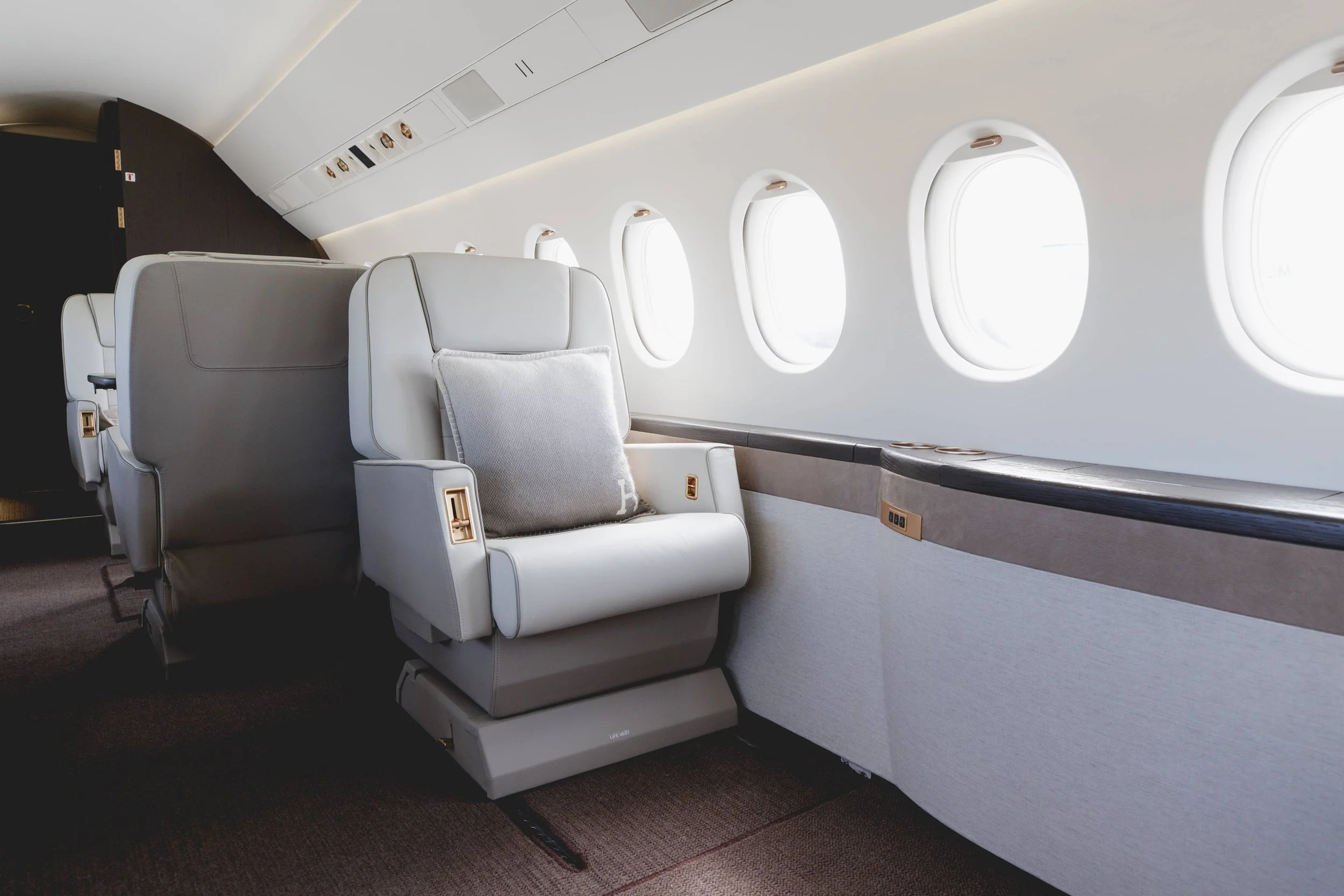 View of airplane interior with white leather seat, gray pillow, and windows along the side of the plane.