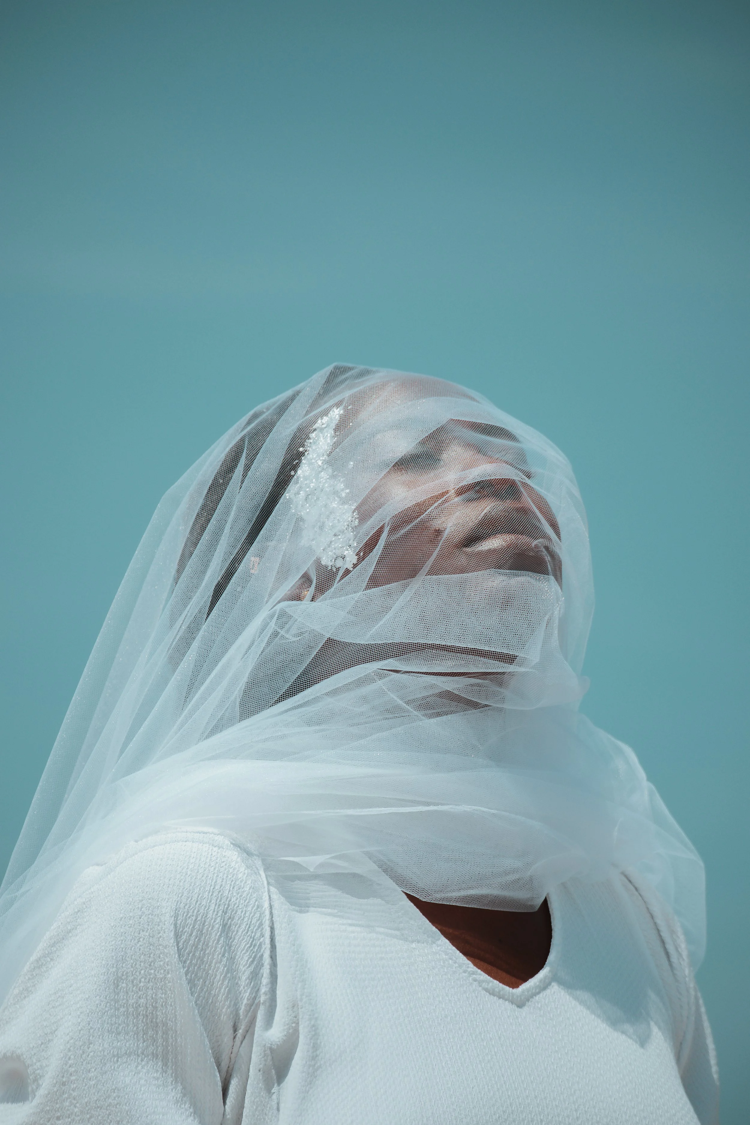 Une personne porte un voile blanc en tissu léger sur la tête et le visage, avec un fond bleu clair.