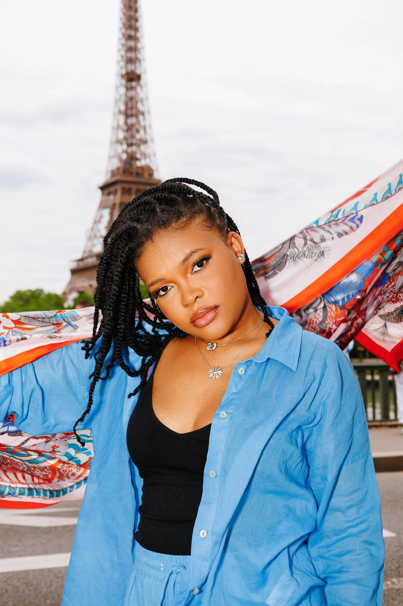 Femme debout devant la Tour Eiffel, portant un haut noir et une chemise bleue, avec un foulard coloré dans le dos.