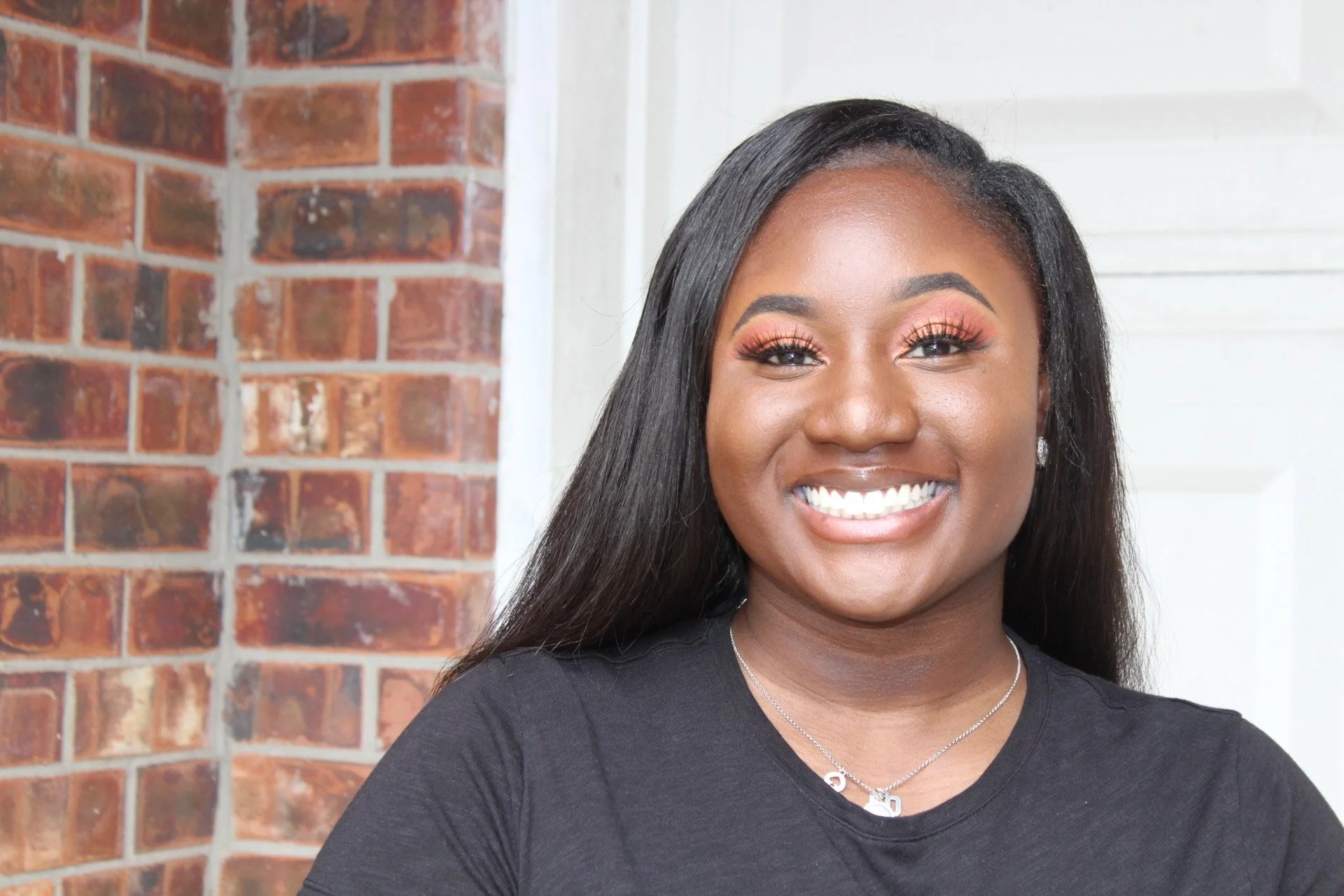 Dr. Akua P. Twumasi, Ed.D, in a black top and a silver necklace, standing in front of a brick wall and white paneling.