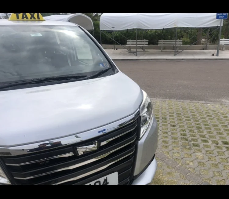 Close-up of the front of a white taxi van parked on a paved surface near a sidewalk and benches, with tents in the background.