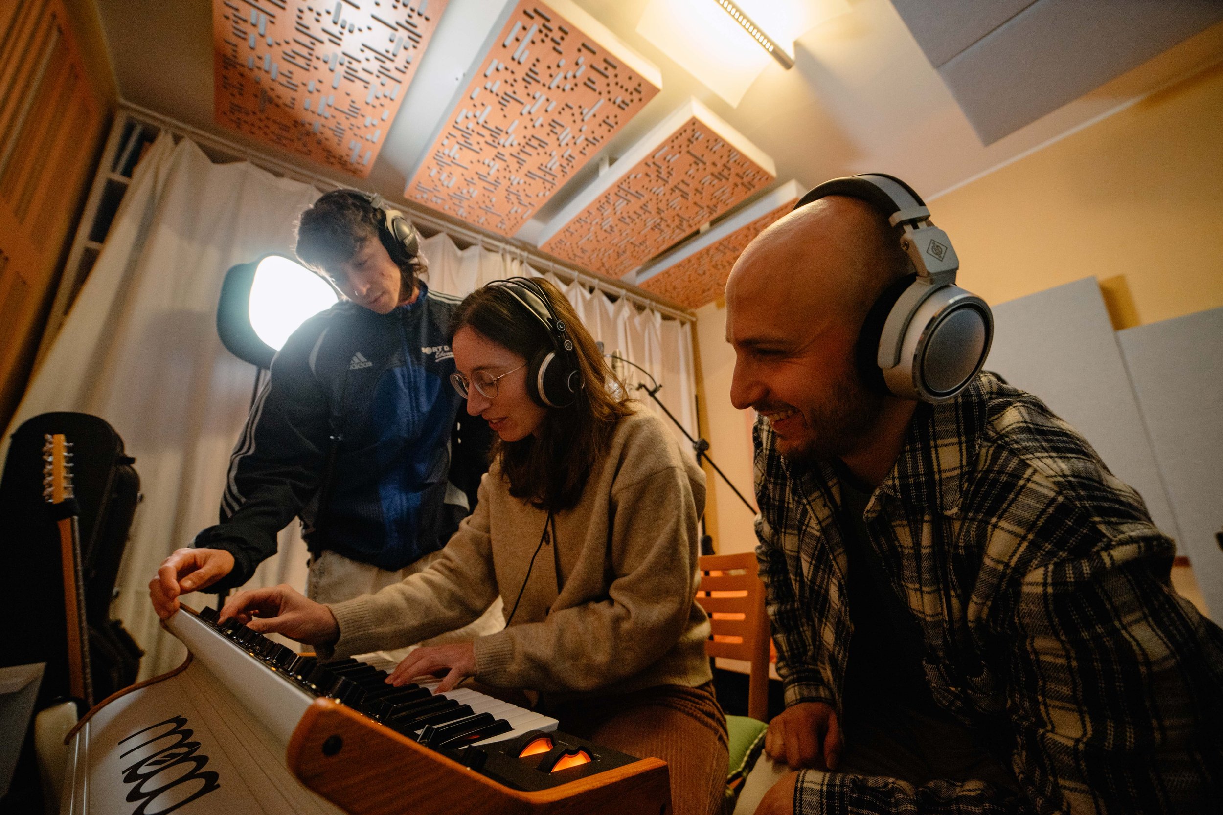 Three people wearing headphones in a music studio, one playing a keyboard, surrounded by musical equipment and soundproof panels.
