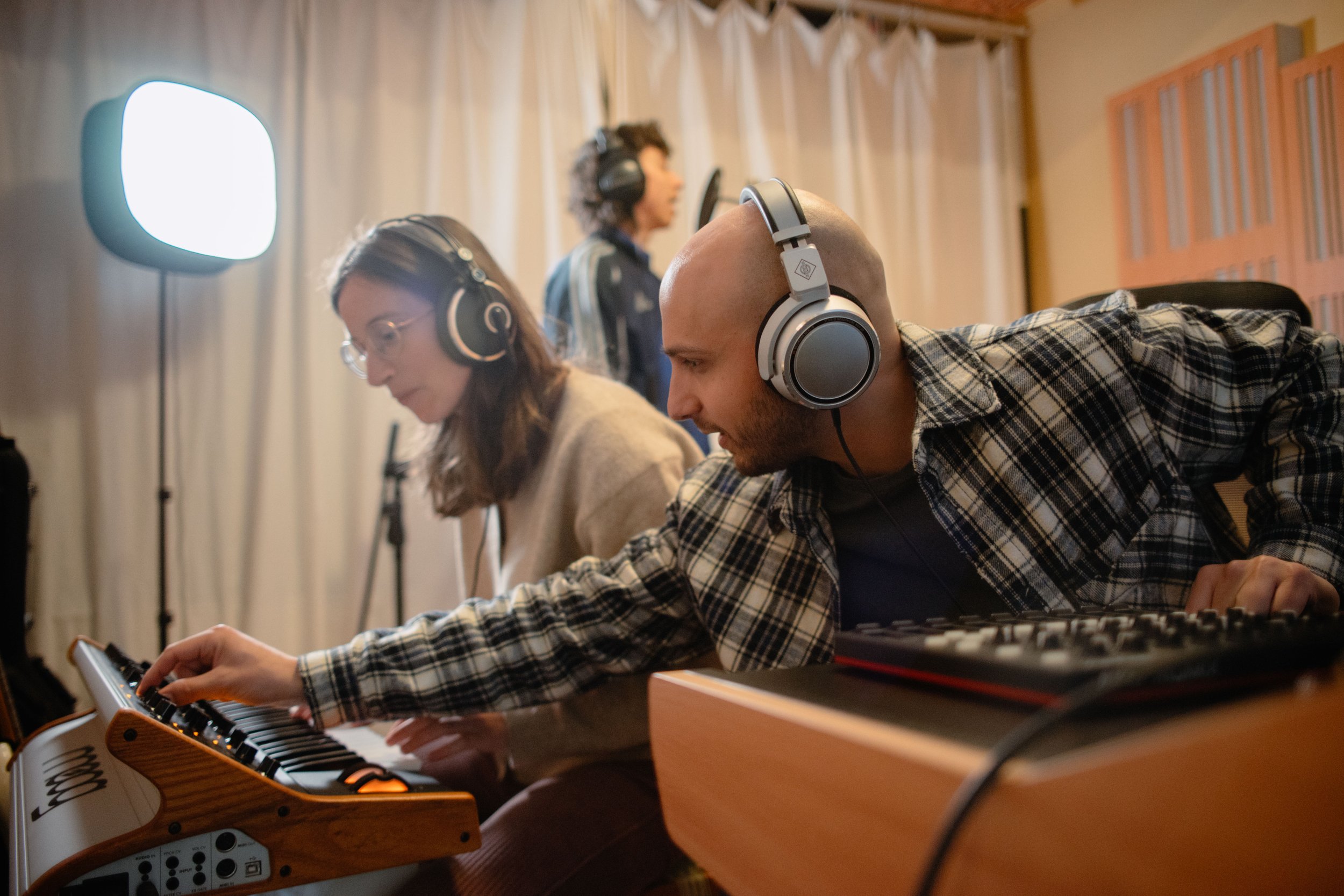 Two people wearing headphones work on music production equipment in a studio, with a third person in the background near a microphone.