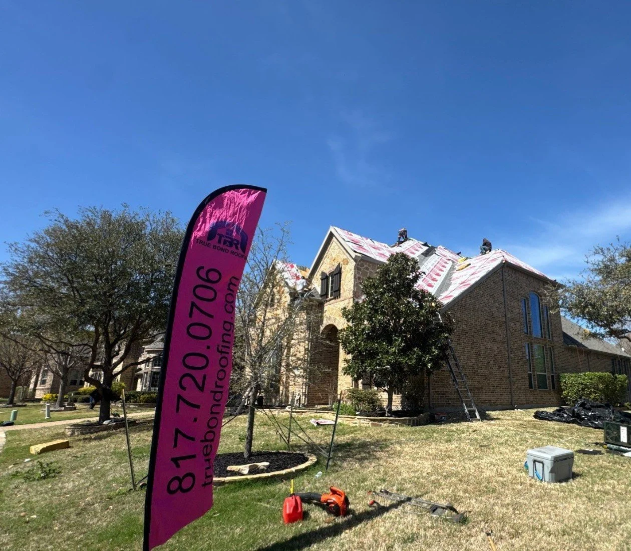 True Bond Roofing crew installing a new residential roof in Dallas-Fort Worth, with roofing underlayment, workers on the roof, and the company's pink jobsite flag displayed.