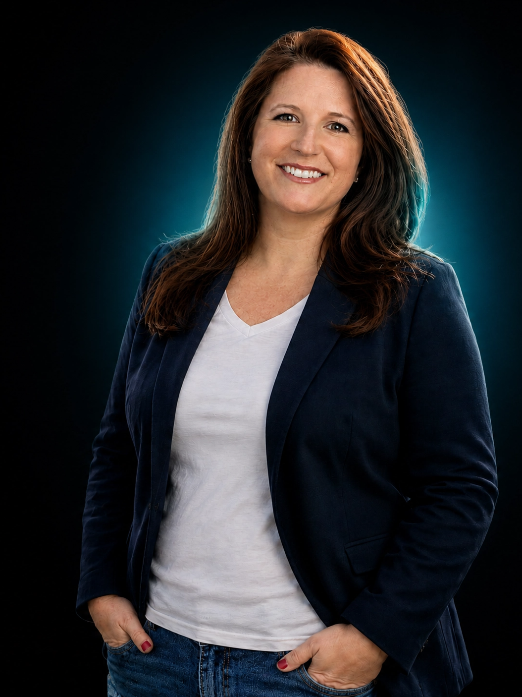 A woman with brown hair, smiling, wearing a navy blazer, white V-neck shirt, and jeans, standing against a dark background with a blue halo effect.