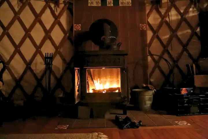 Cozy indoor scene with a burning fireplace in a yurt, surrounded by tiled walls and wooden lattice design. Fireplace tools and a metal bucket are nearby on the floor.