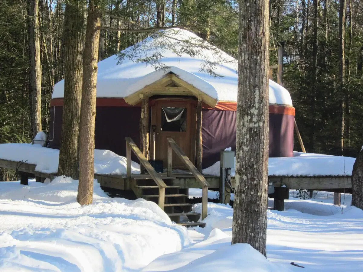 Snow-covered yurt in snowy forest setting with wooden steps and surrounding trees.