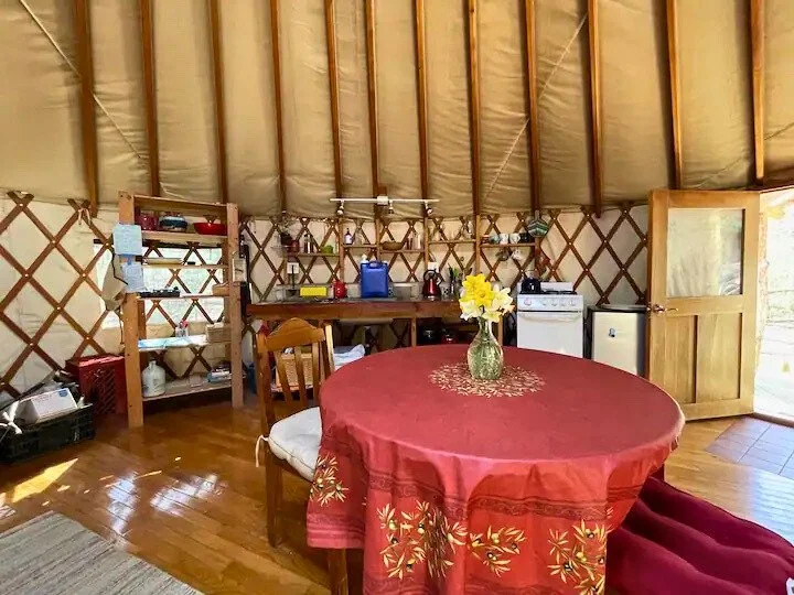 Interior of a yurt with a red tablecloth-covered table at center, a vase with yellow flowers, wooden shelves with kitchen items, a chair, a small fridge, and a stove.