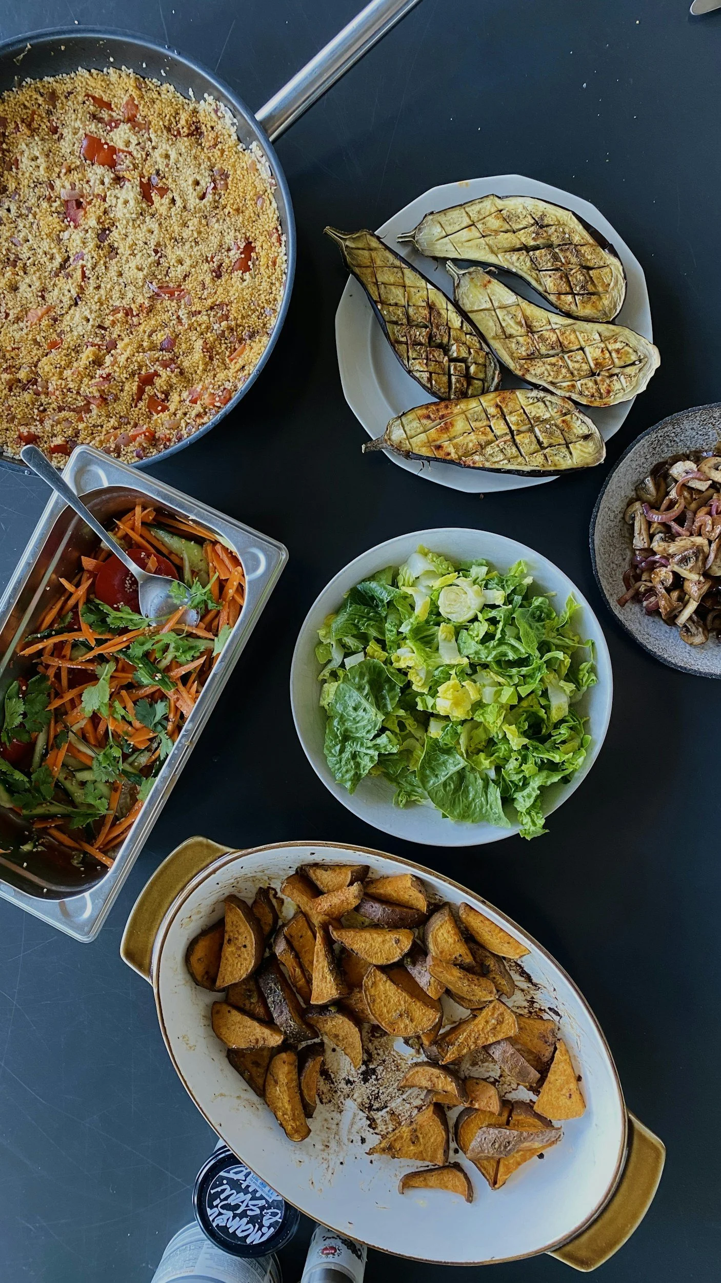 A variety of cooked dishes on a black table, including grilled eggplants, a bowl of mixed green salad, roasted sweet potatoes, a vegetable quiche, a fresh vegetable salad, and a stir-fried mushroom dish.