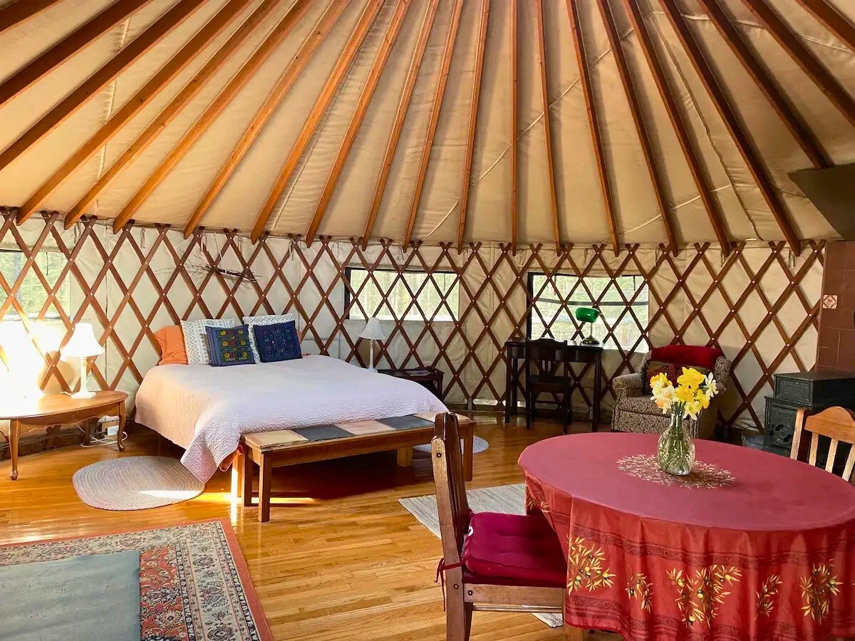 Interior of a yurt with a bed, round dining table, chairs, and a small desk. The wooden lattice structure and supporting beams are visible. Sunlight illuminates the space, creating a cozy atmosphere with colorful textiles and a vase of flowers on the