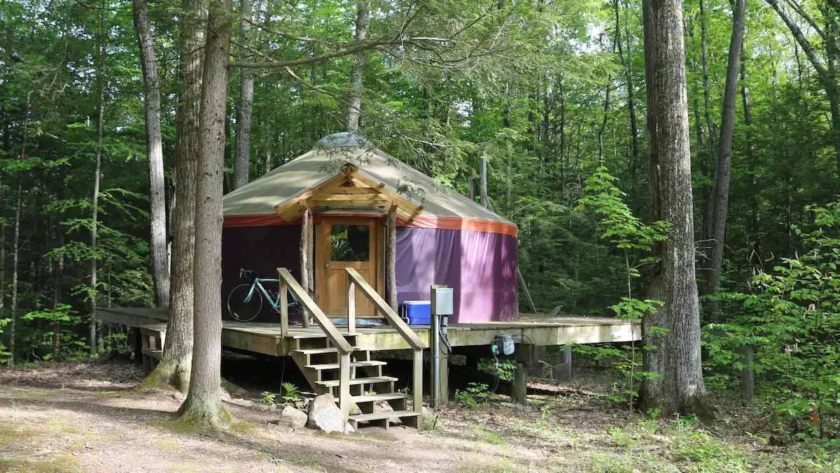 A yurt with a wooden deck surrounded by trees in a forest. A bicycle and some items are on the deck.