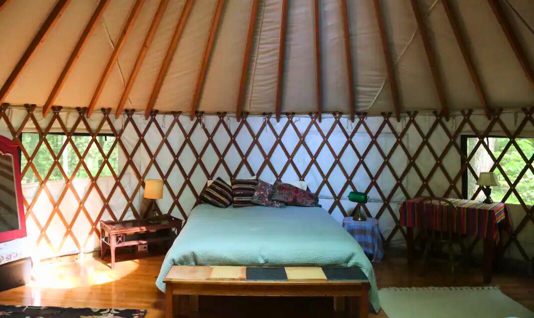 Interior of a yurt with a bed, wooden frame, and colorful pillows, surrounded by lattice walls. There are small tables with lamps and a window overlooking a forested area.