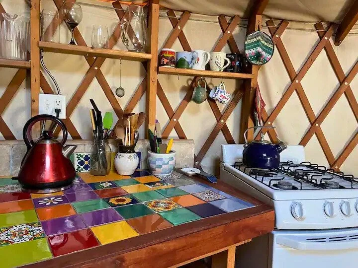 Cozy kitchen in a yurt with colorful tile countertop, wooden shelves holding mugs and glasses, a red kettle on the counter, and a blue kettle on a white gas stove.