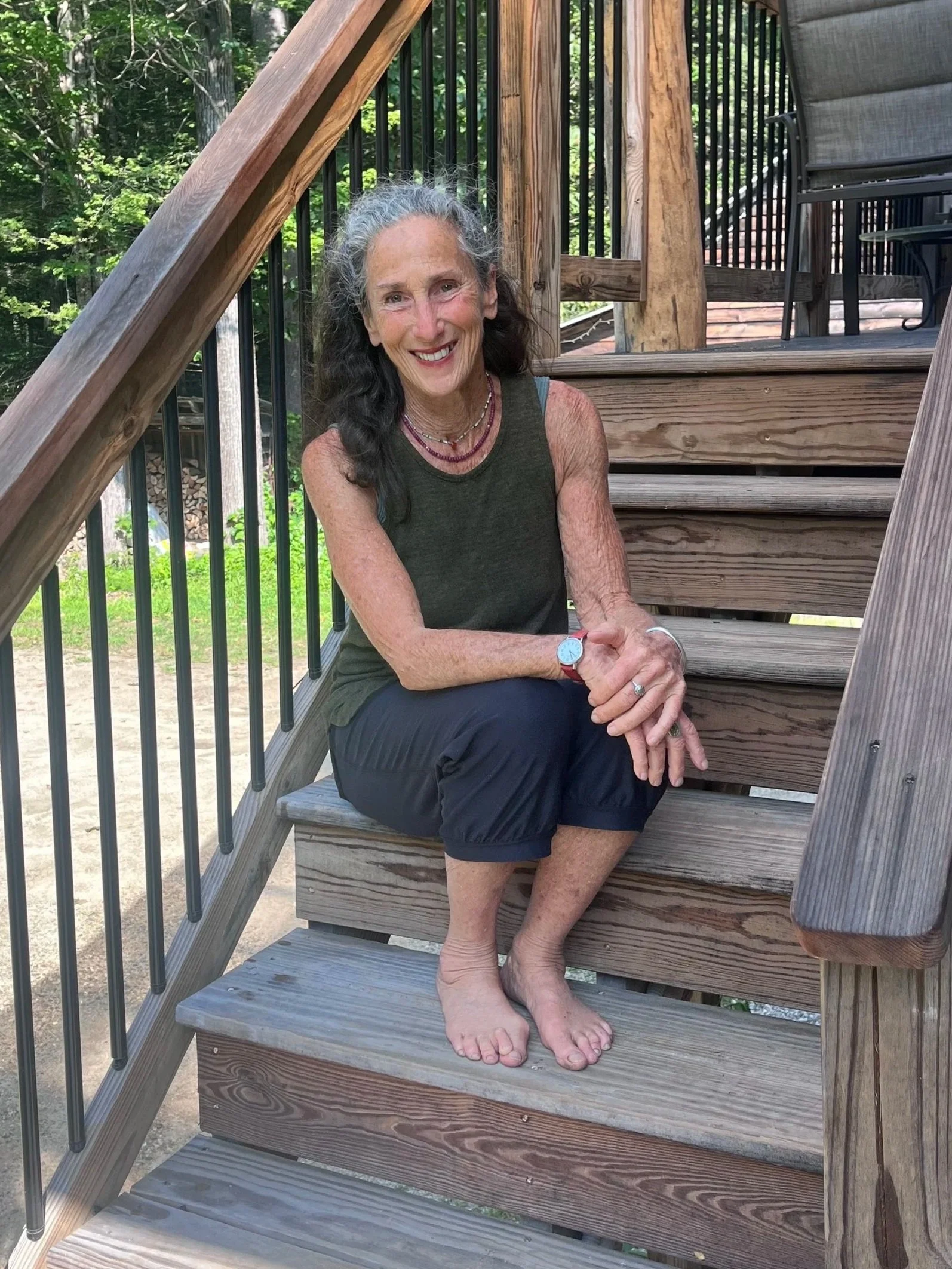 A woman with gray and black hair sitting on wooden stairs outdoors, smiling, wearing a green sleeveless top, black pants, and jewelry, with trees in the background.