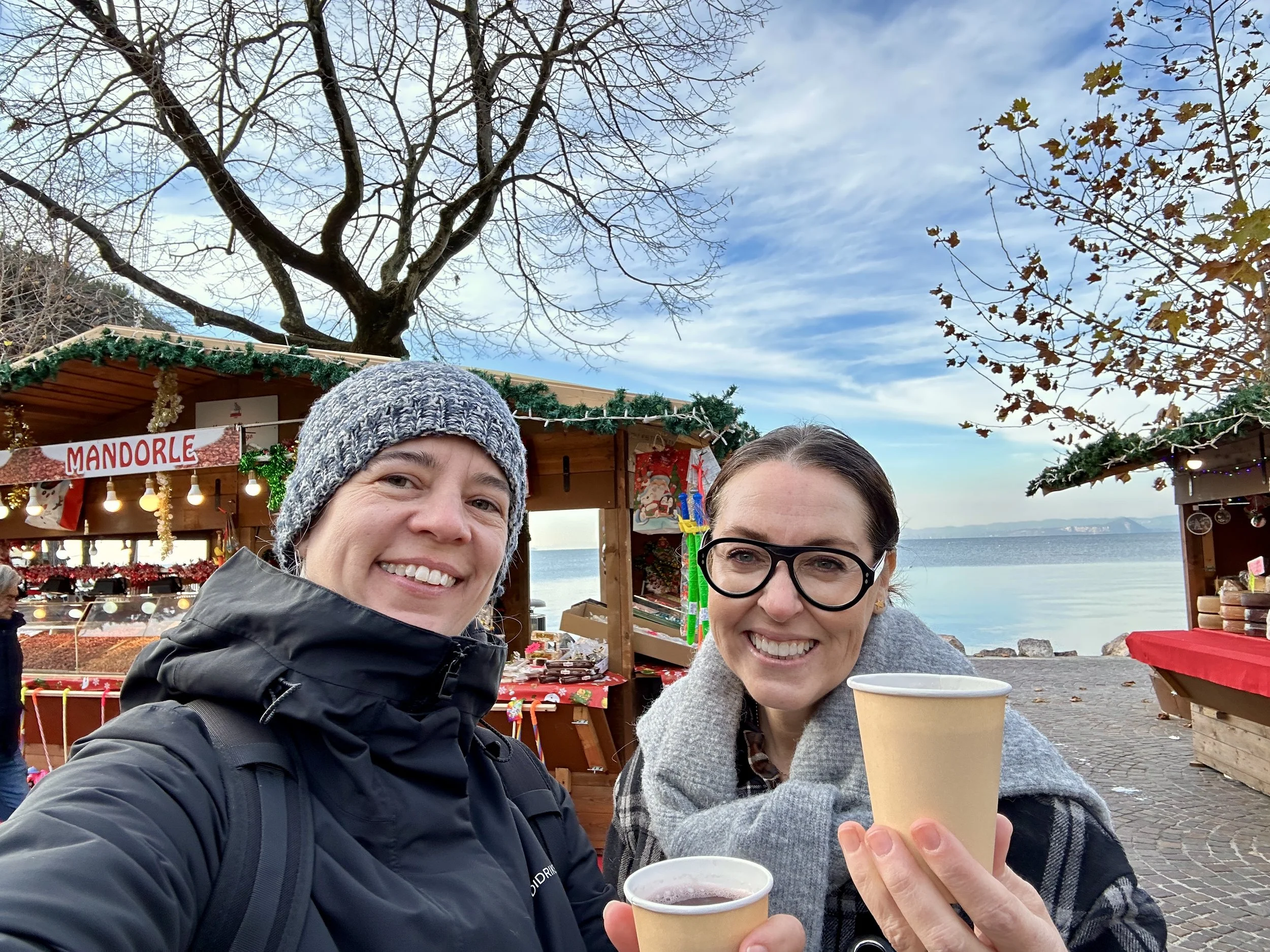 Two women holding hot drinks and smiling in front of a Christmas market stall beside Lake Garda with bright blue skies