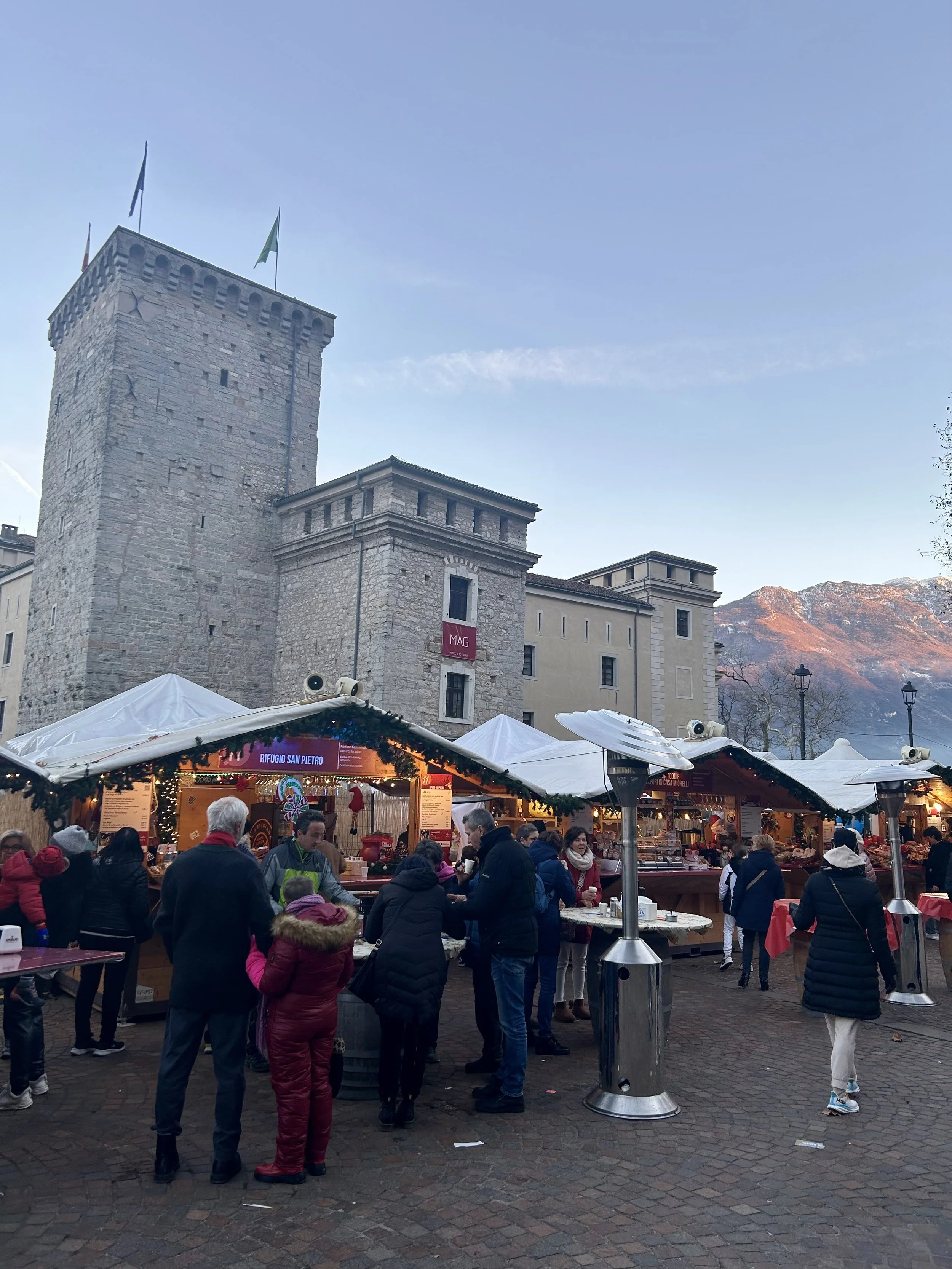 Outdoor Christmas market beside a historic stone tower with mountain peaks glowing pink in the distance at sunset.