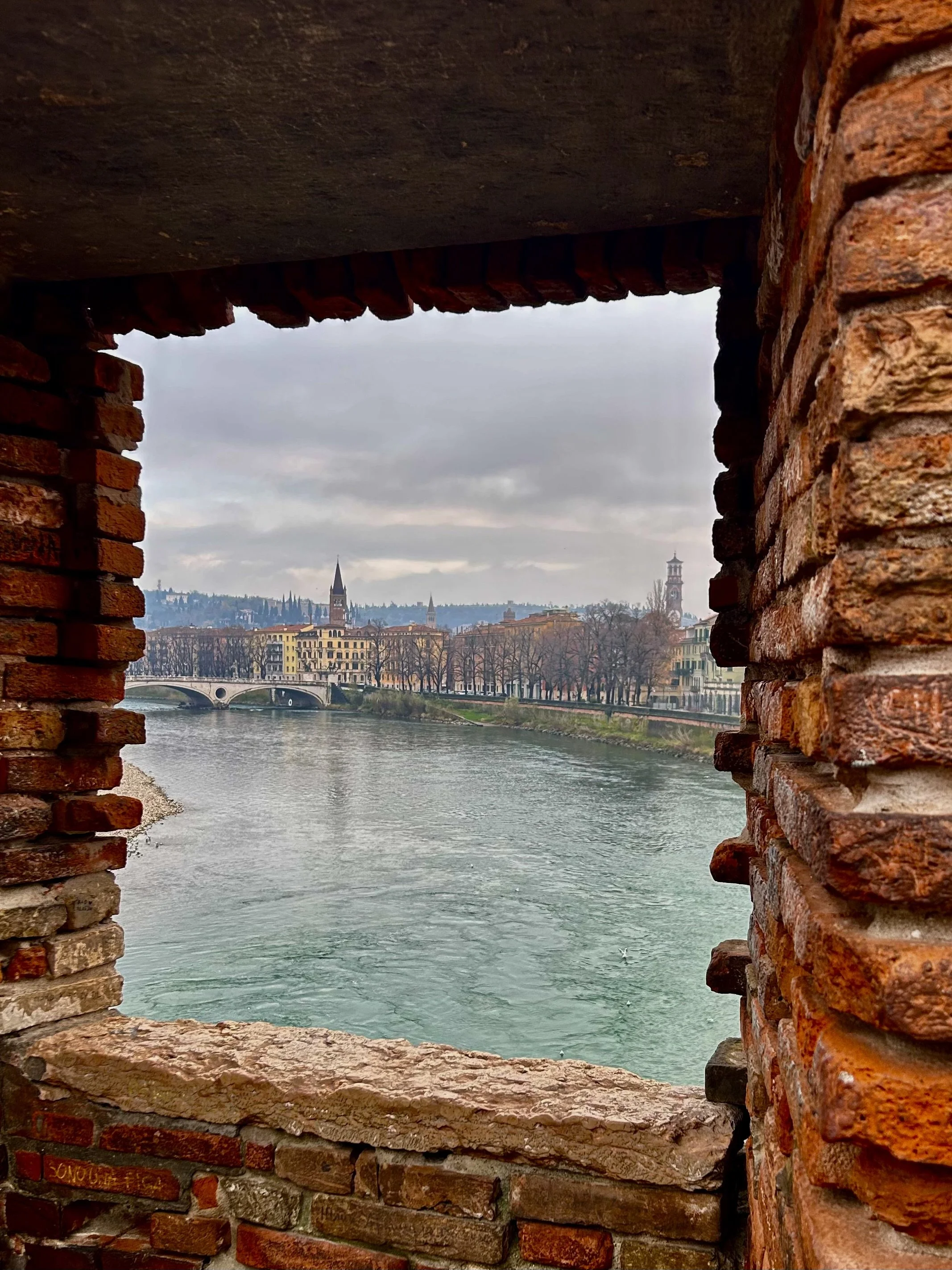 “View of the Adige River and Verona’s skyline framed through the brick window of Castelvecchio, on a cloudy winter day.”